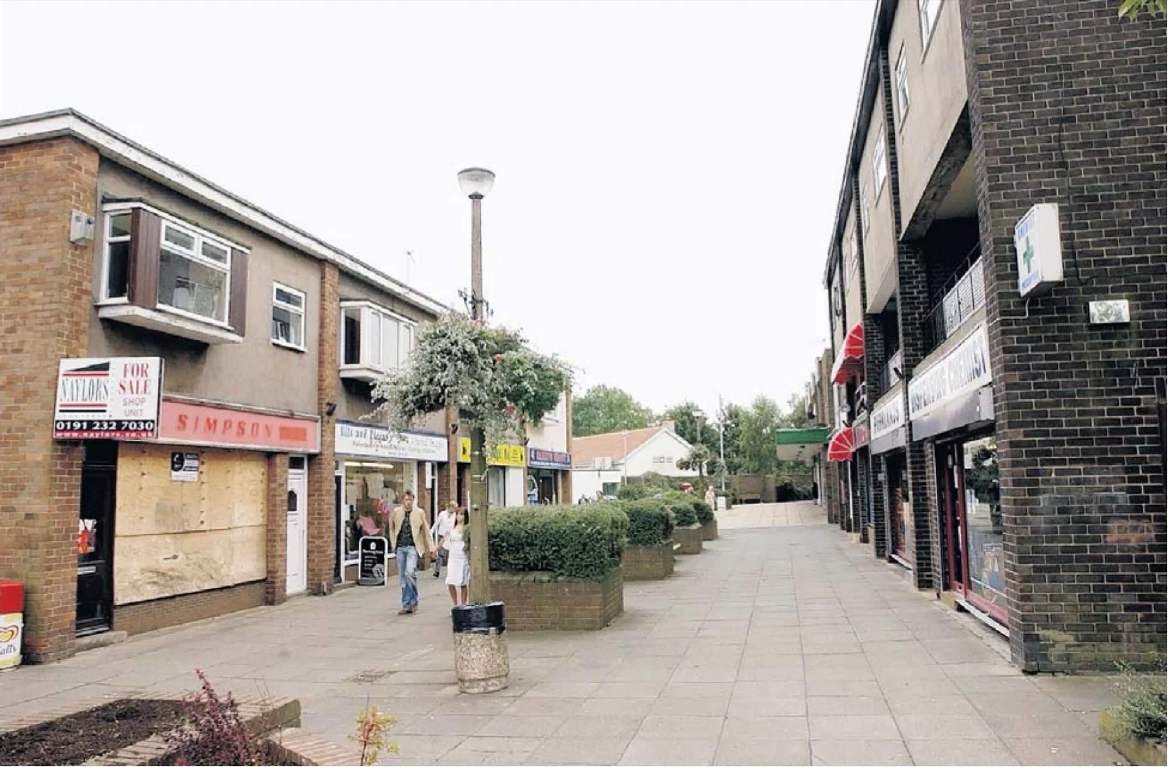 A 1960s pedestrian shopping precinct with flat-roofed brick units, a concrete lamp post and planters, one shopfront boarded up