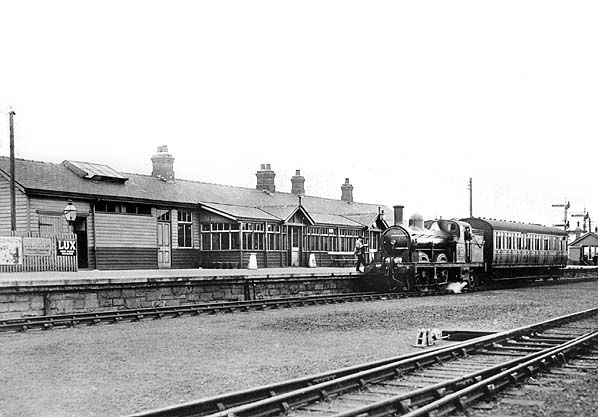 Black and white photograph of a steam locomotive and carriages standing at a railway station platform with a long timber station building
