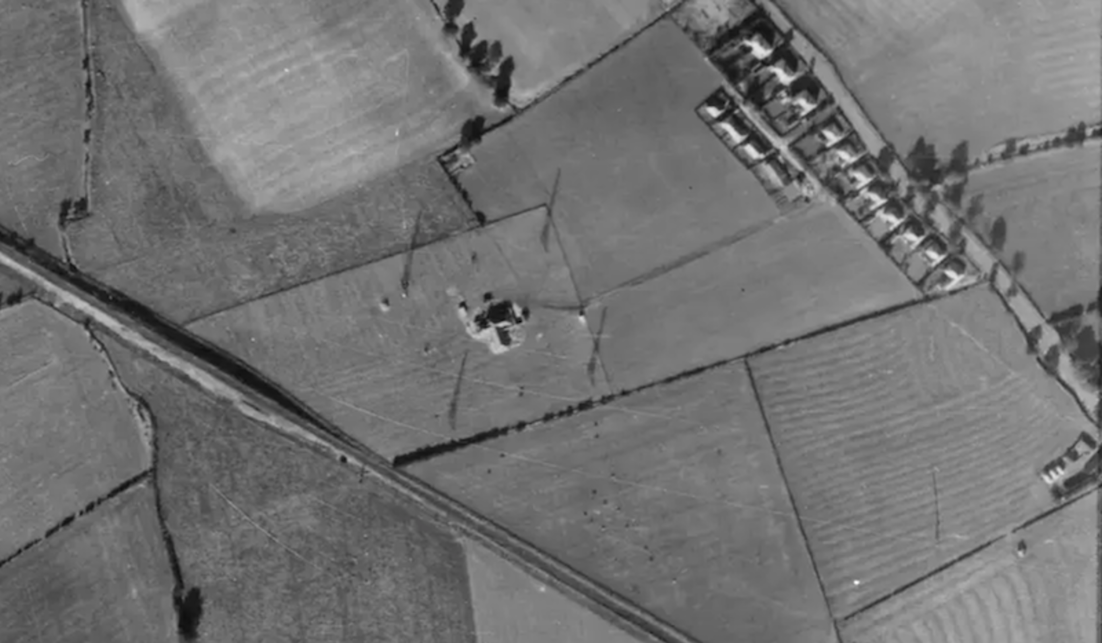Black and white aerial photograph of farmland with a small circular structure and radiating tracks in the centre of a field