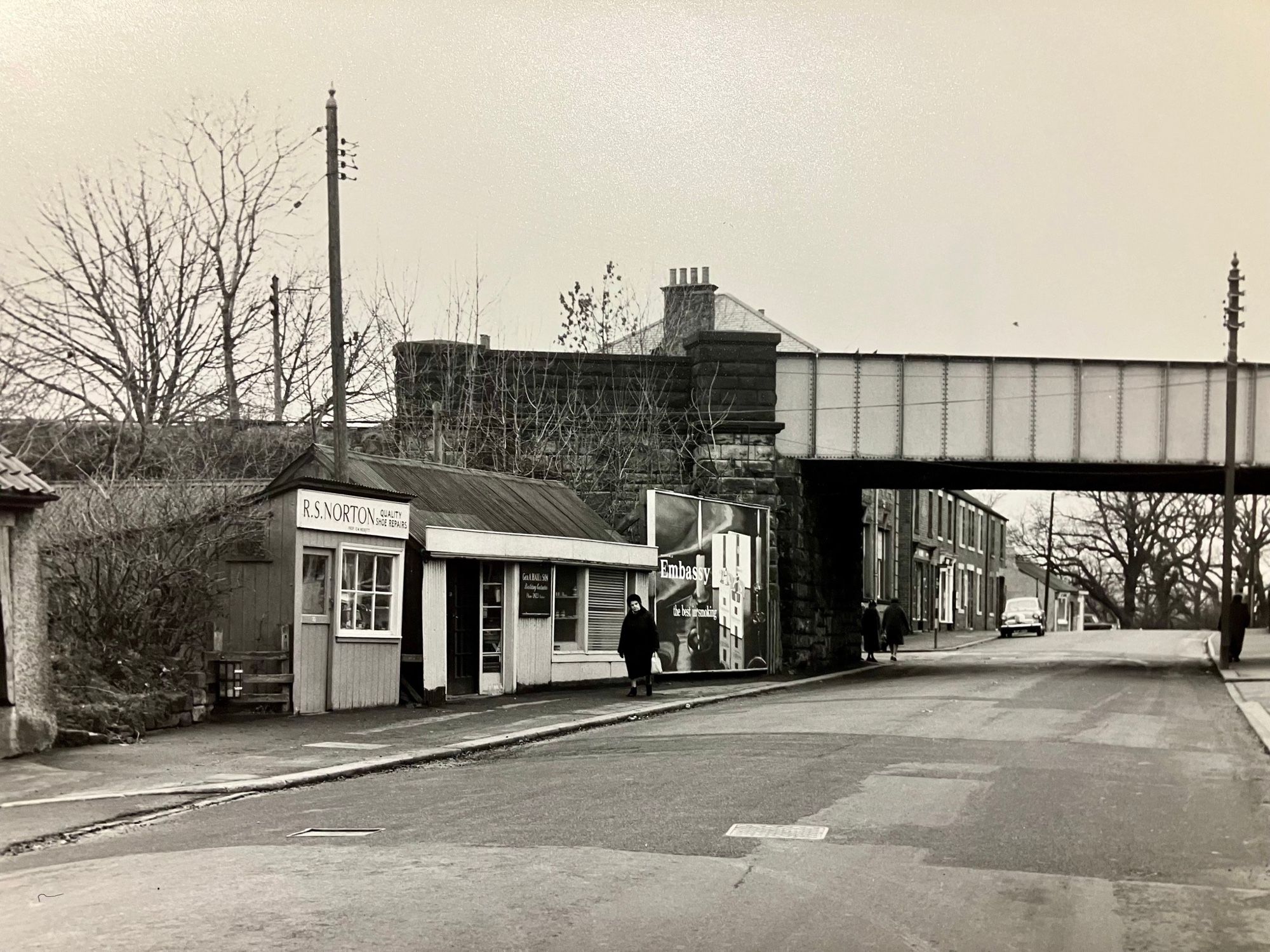 Black and white photograph of single-storey shops beside a road, with a steel railway bridge crossing overhead