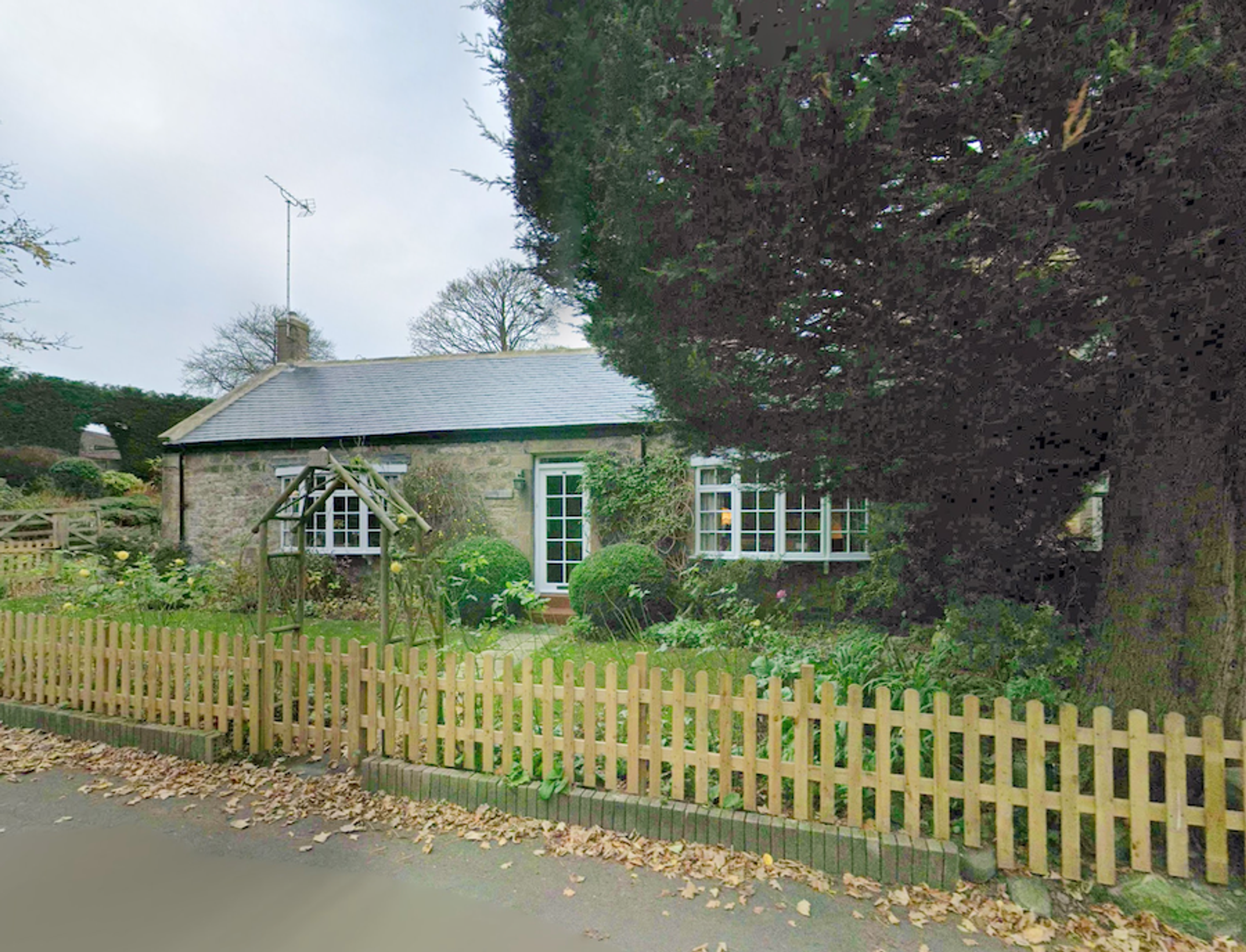A stone cottage with a slate roof and gabled porch behind a wooden picket fence, with a large tree overhanging the garden