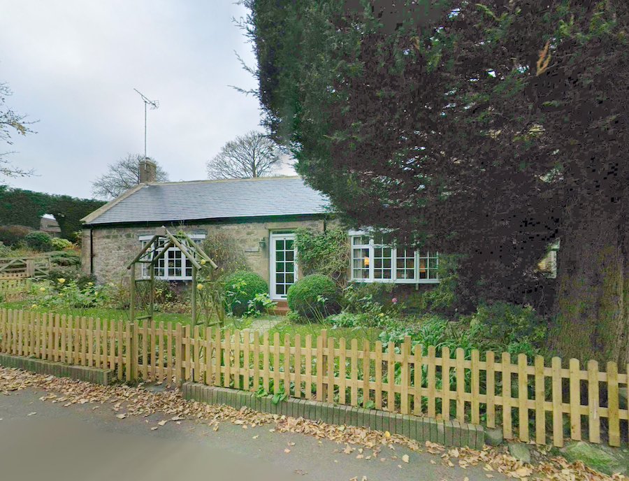 A stone cottage with a slate roof and gabled porch behind a wooden picket fence, with a large tree overhanging the garden