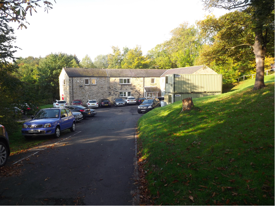 A converted stone mill building with a modern green extension, cars parked on the sloping drive beneath autumn trees