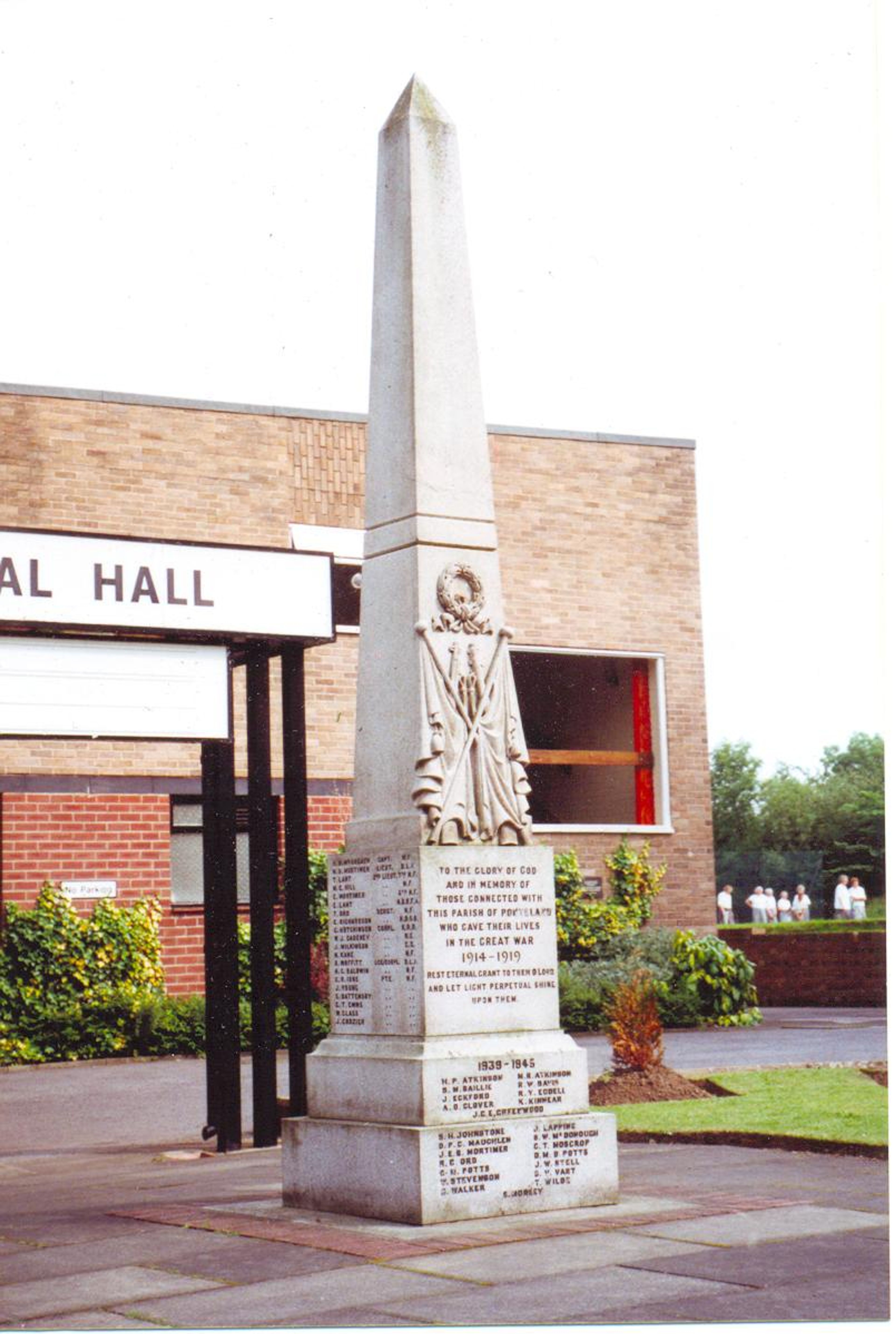 A tall stone obelisk war memorial carved with a soldier and inscribed name panels, standing outside a brick village hall