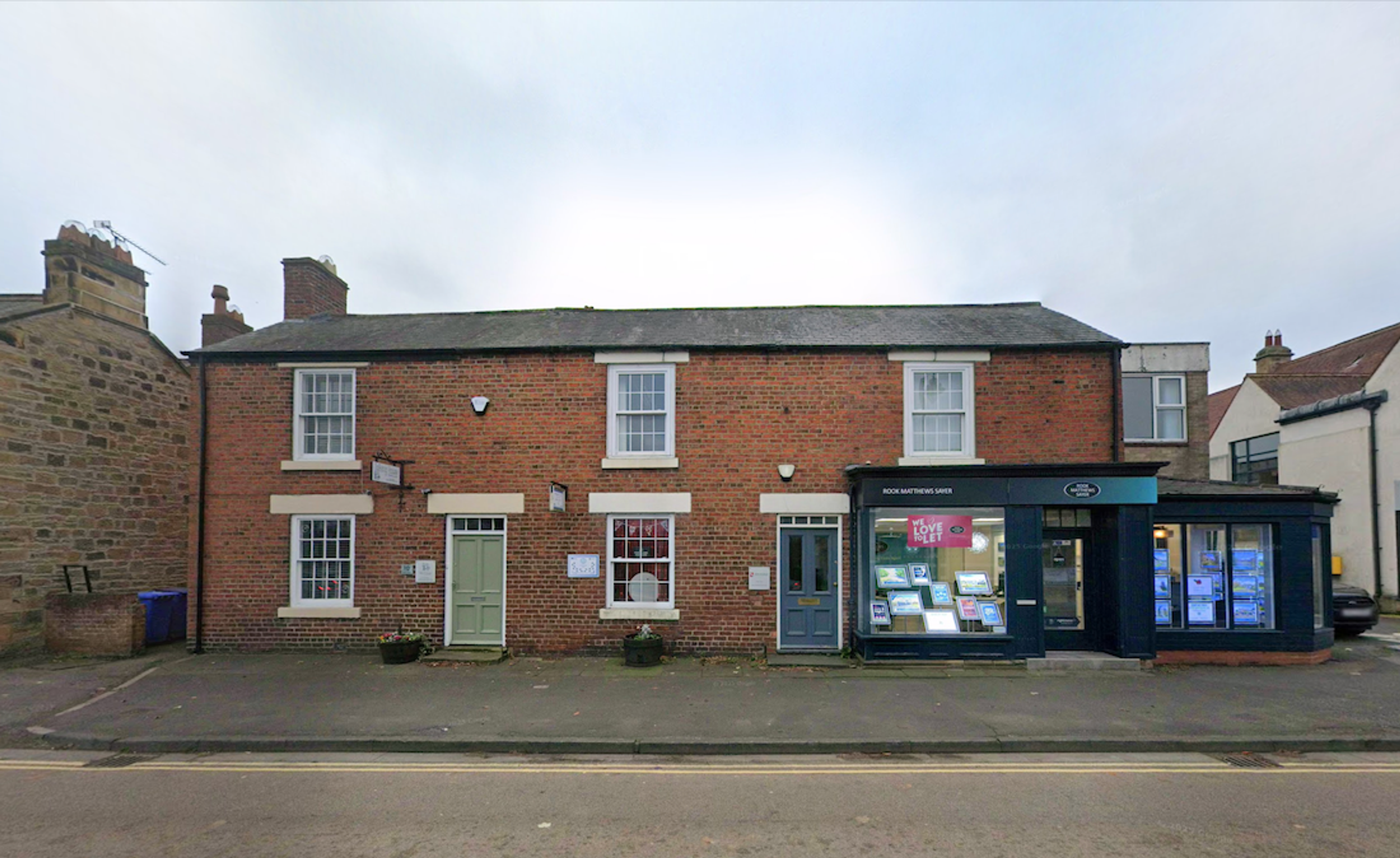 A red brick house and adjoining shopfront with sash windows and a painted bay window, standing on the roadside