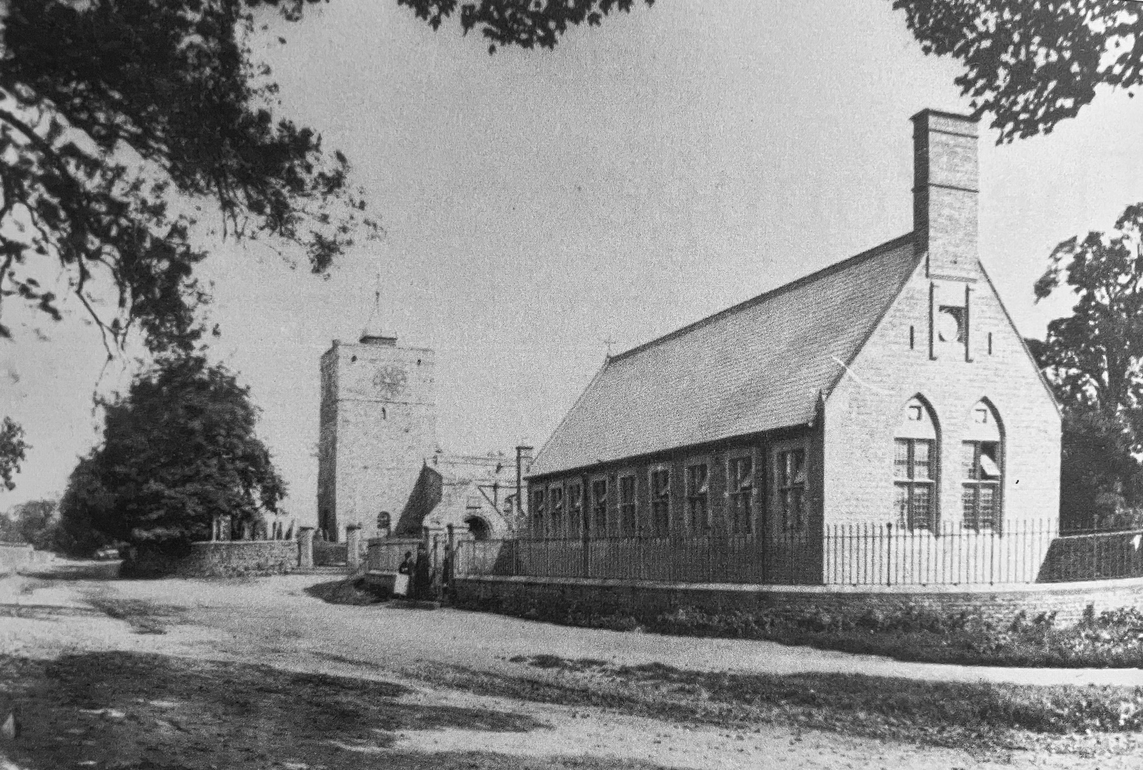 Black and white photograph of a stone school building with tall gabled windows and a chimney, a church tower beyond