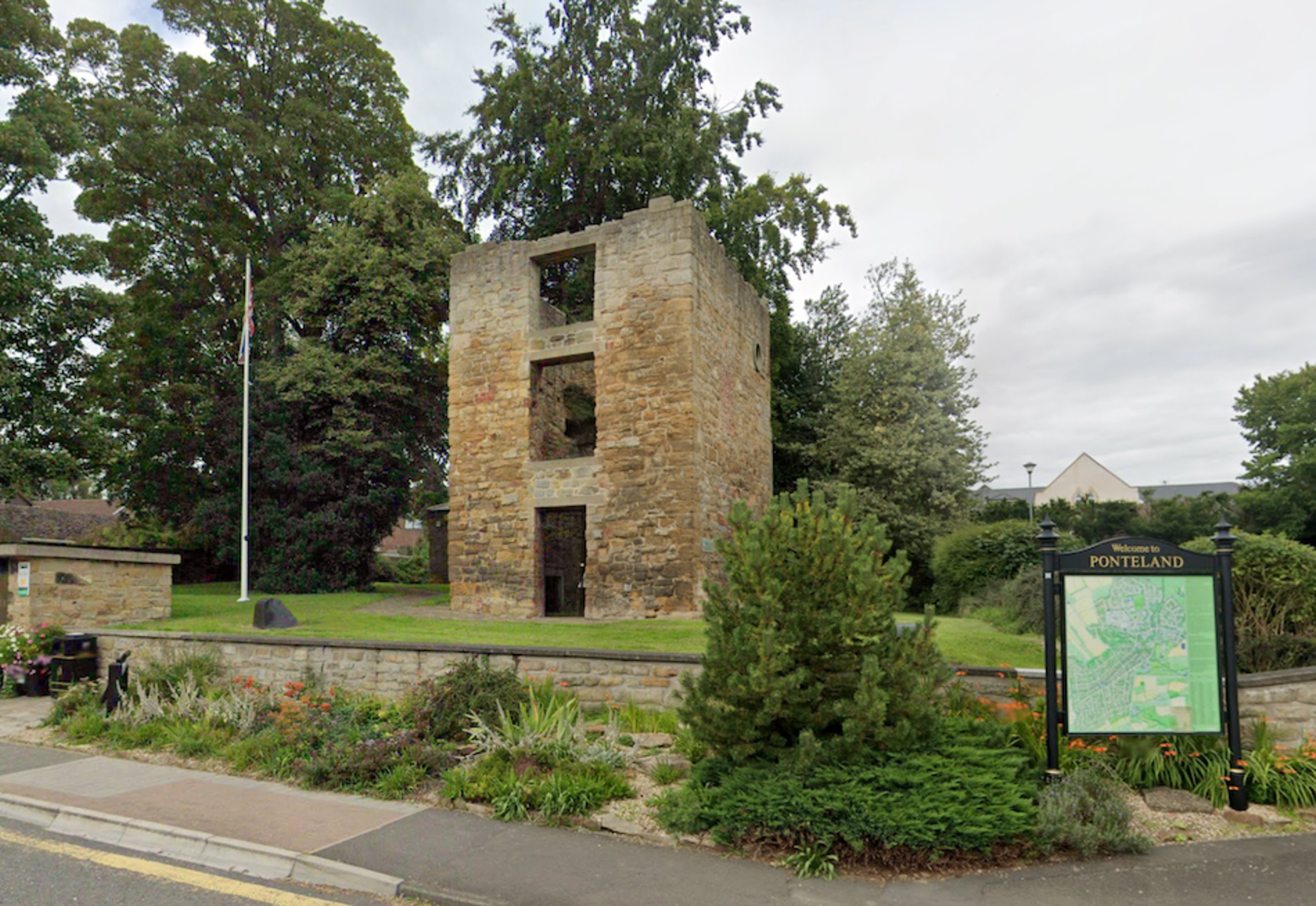 The roofless stone shell of a medieval tower standing on a lawn beside a road, with a village information map board in front