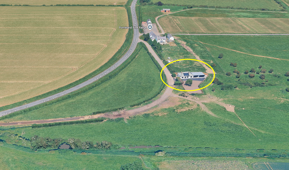 Aerial view of farmland with a yellow ellipse marking a small building beside a track, a main road crossing the fields