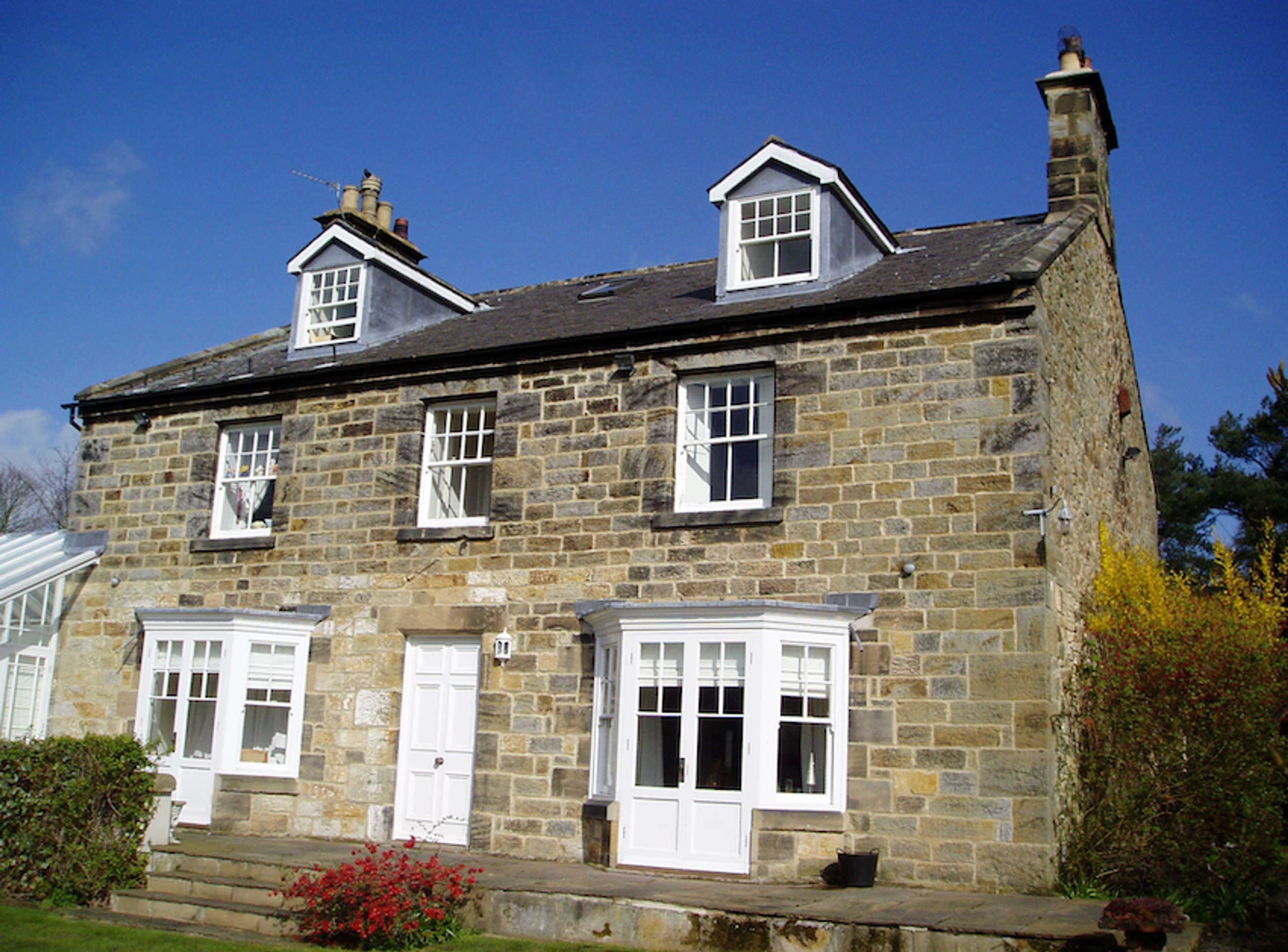 A two-storey stone farmhouse with dormer windows, white sash windows and a central door, in bright sunshine