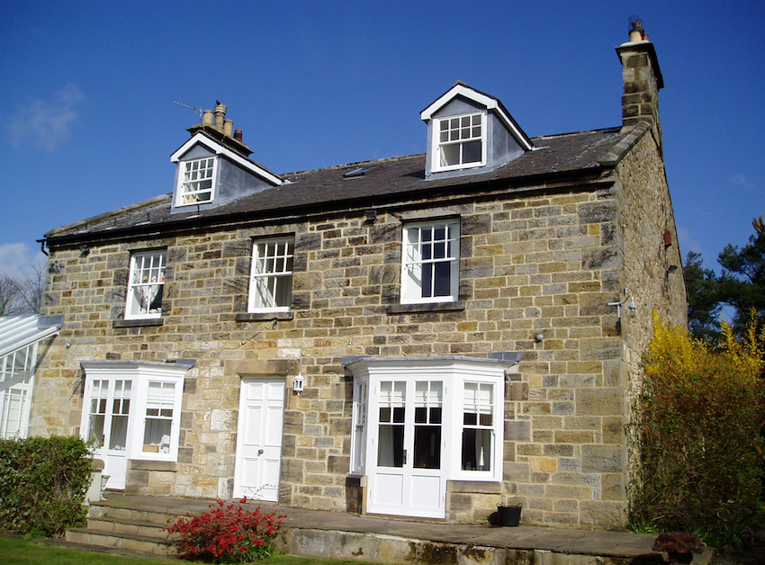 A two-storey stone farmhouse with dormer windows, white sash windows and a central door, in bright sunshine