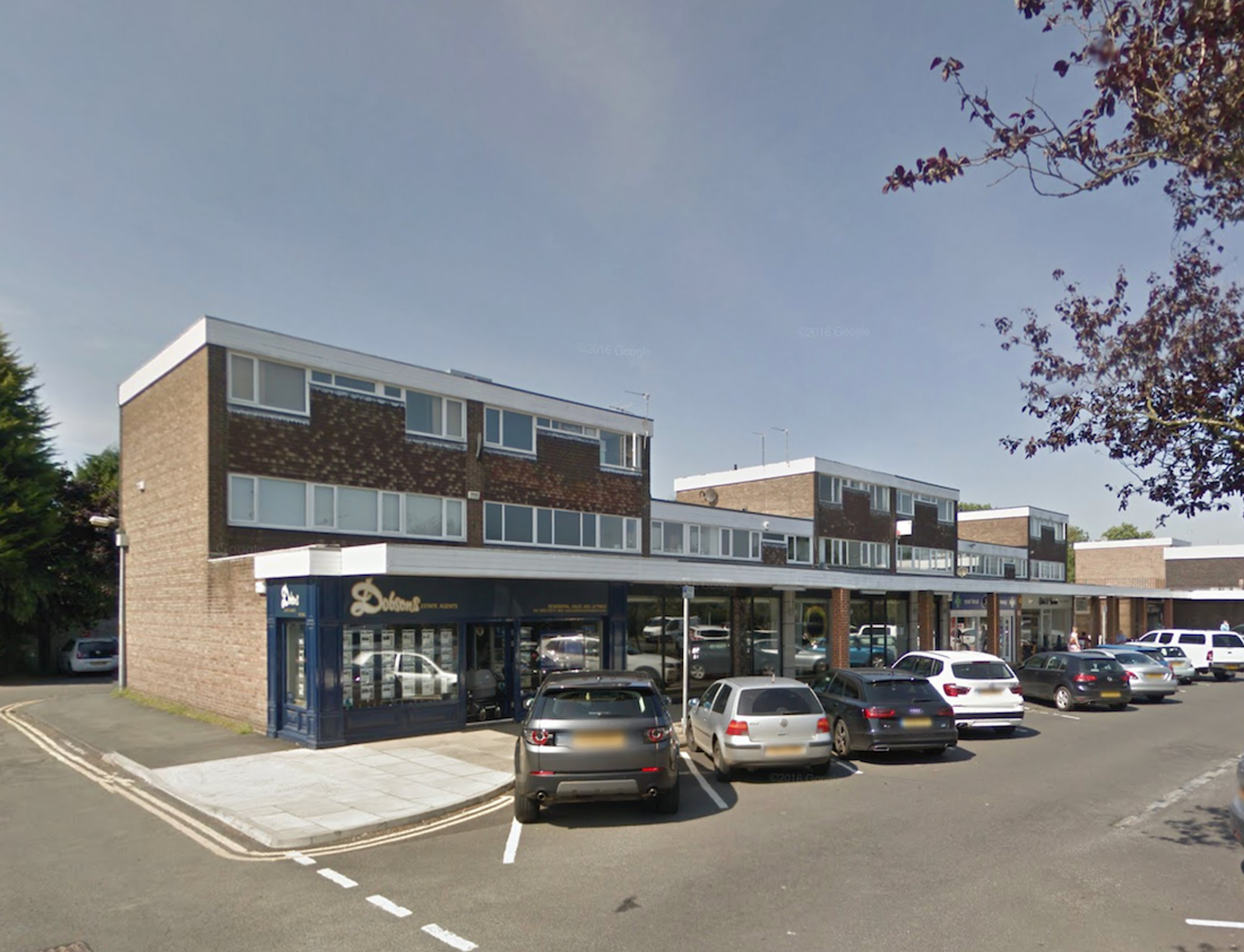 A 1960s parade of shops with flats above, flat-roofed and faced in brick and mosaic panels, cars parked in front