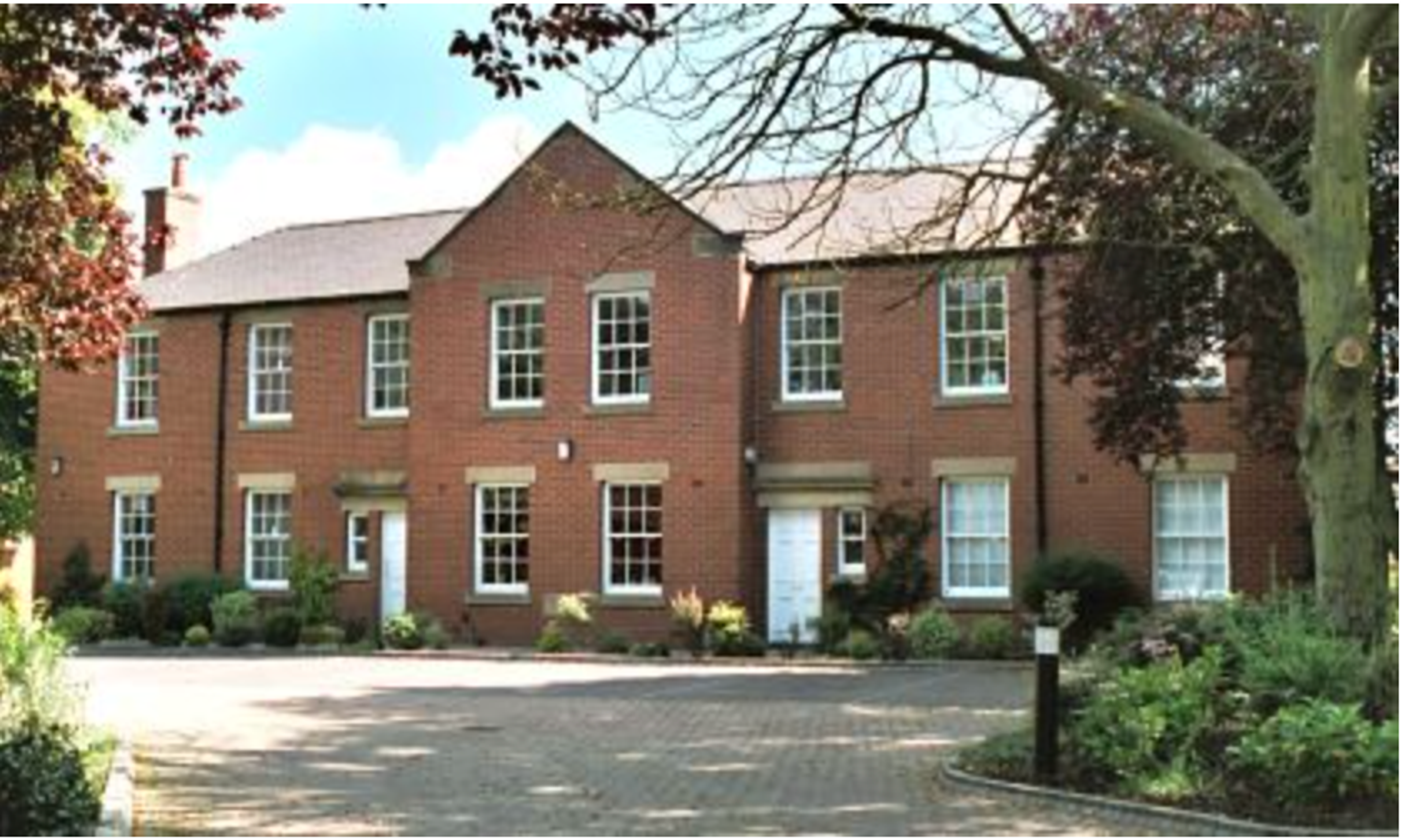A large red brick house with white sash windows and two front doors, behind a block-paved drive