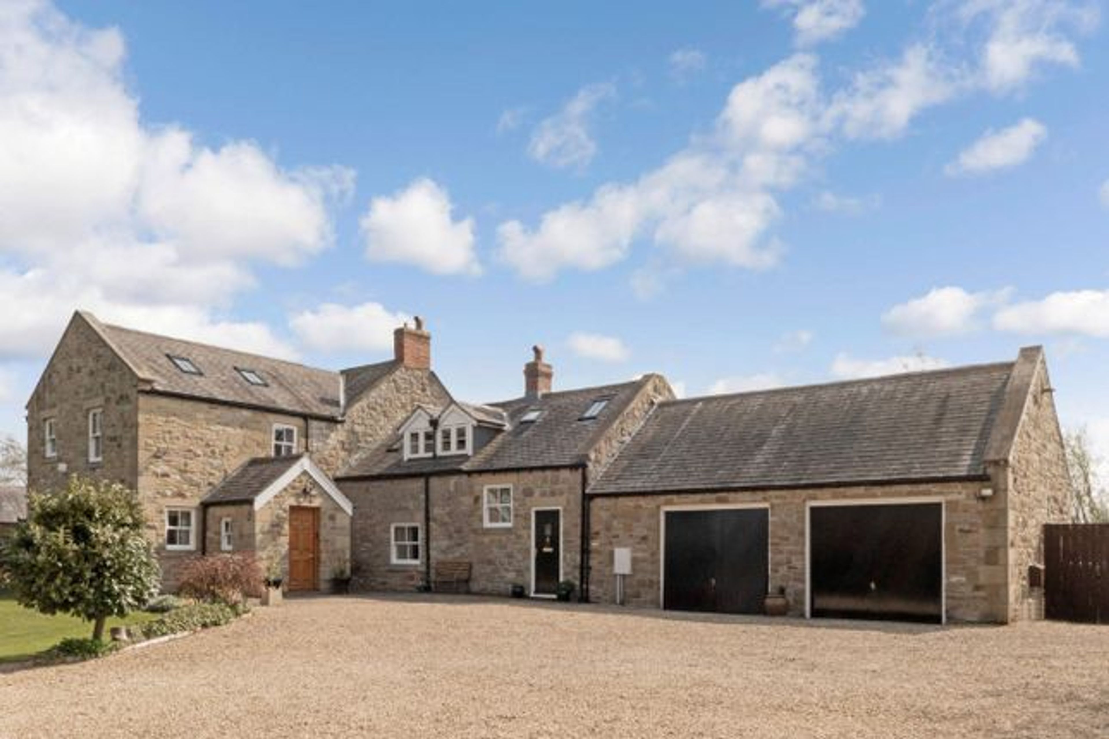 A converted stone farmhouse and attached barn with a gravel courtyard, dormer windows and garage doors, under a blue sky