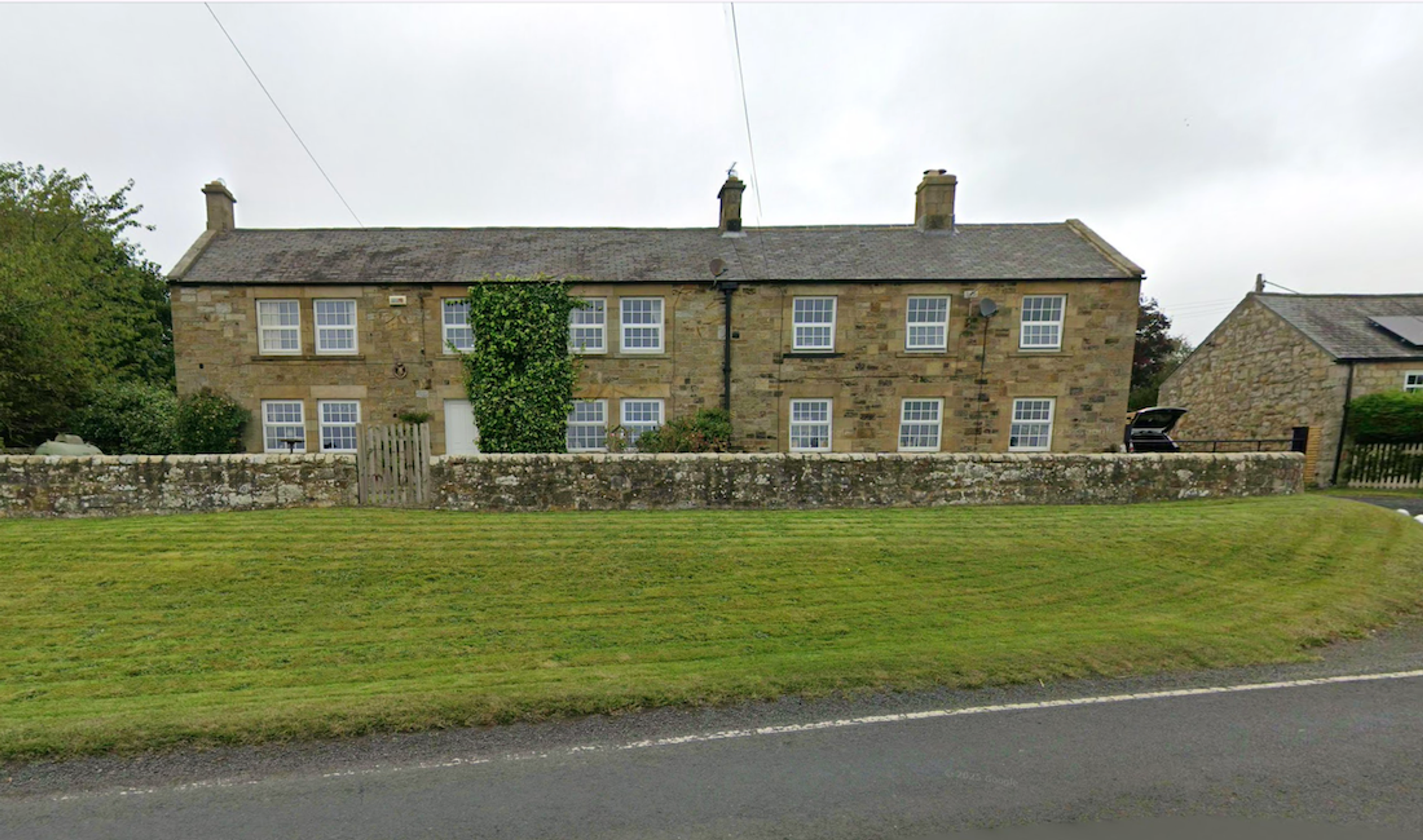 A two-storey stone farmhouse with sash windows and two chimneys, set behind a low stone wall with a grass verge in front