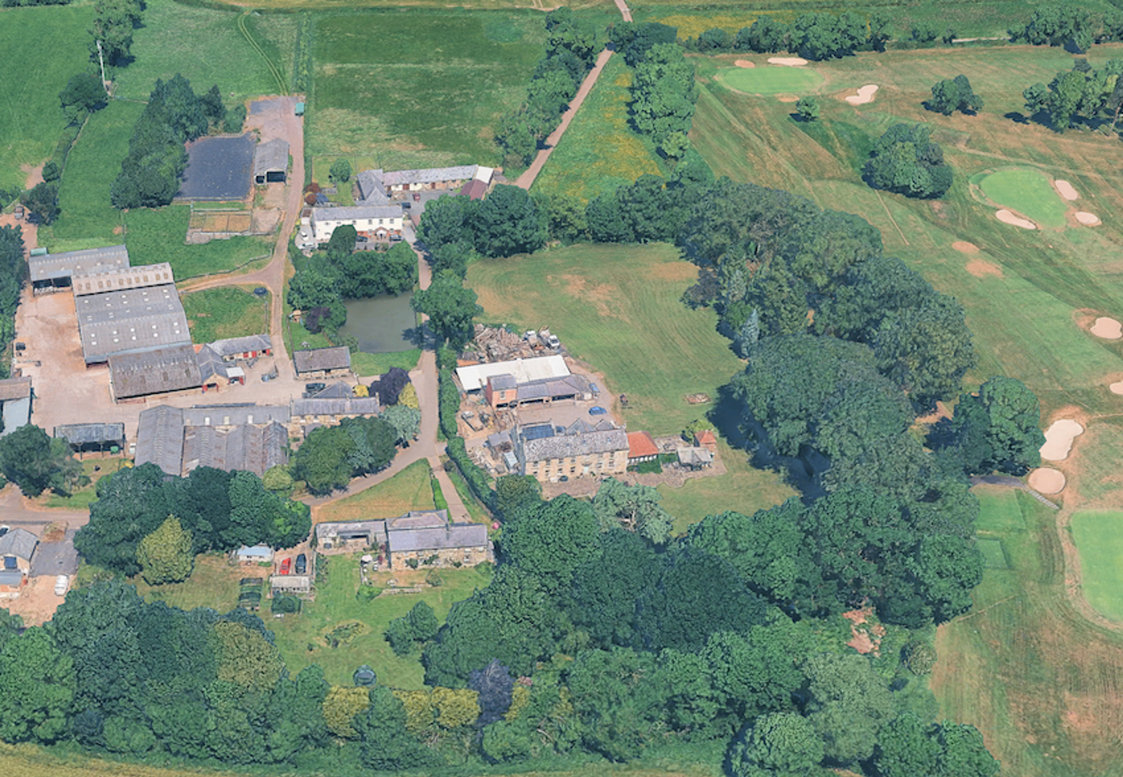 Aerial view of a farmstead with a large house, ranges of outbuildings and a pond, with a golf course alongside