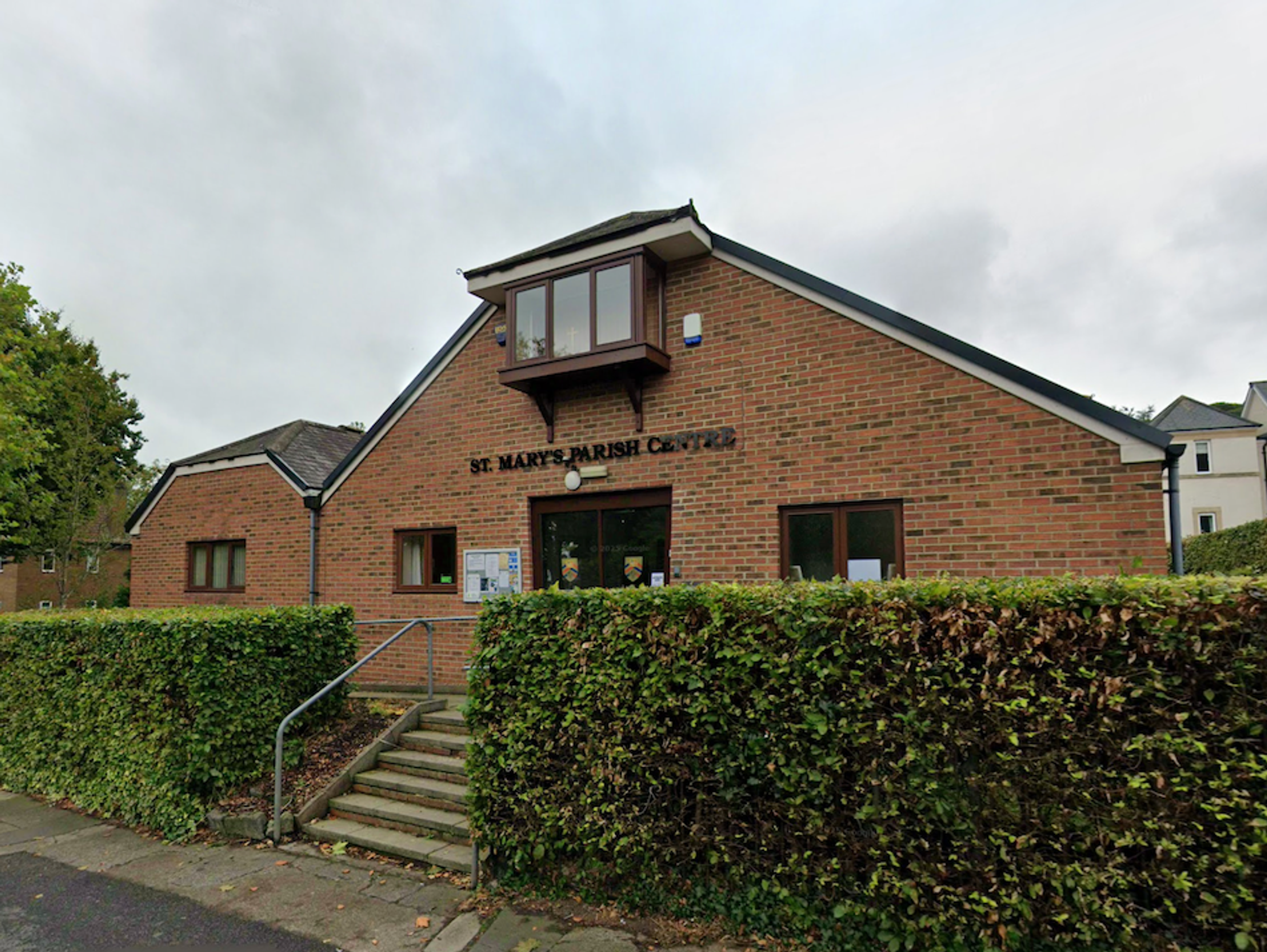 A modern red brick parish centre with a steeply pitched roof and an oriel window, behind a clipped hedge
