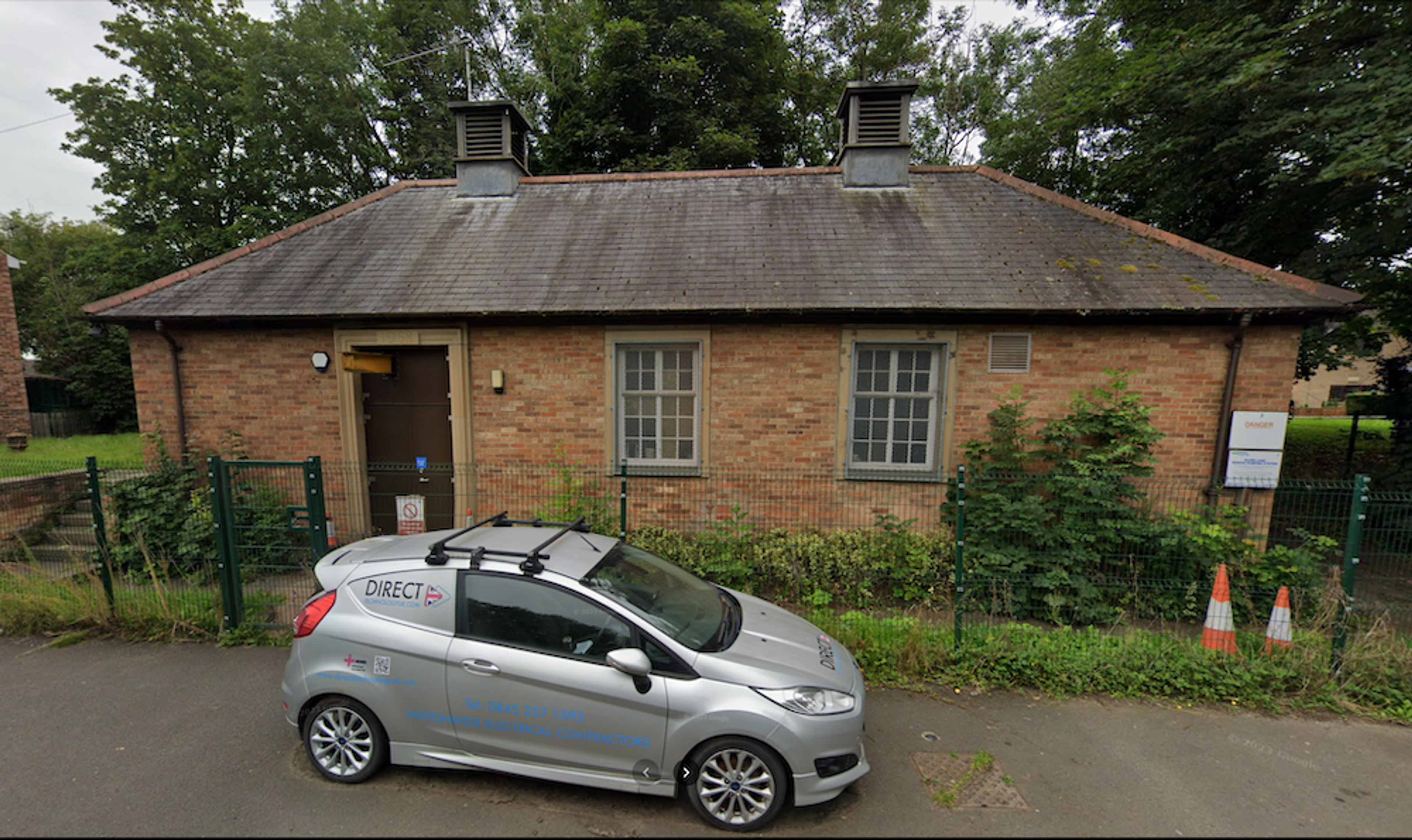 A small single-storey brick building with roof ventilators and a slate roof, behind metal railings with a car parked outside