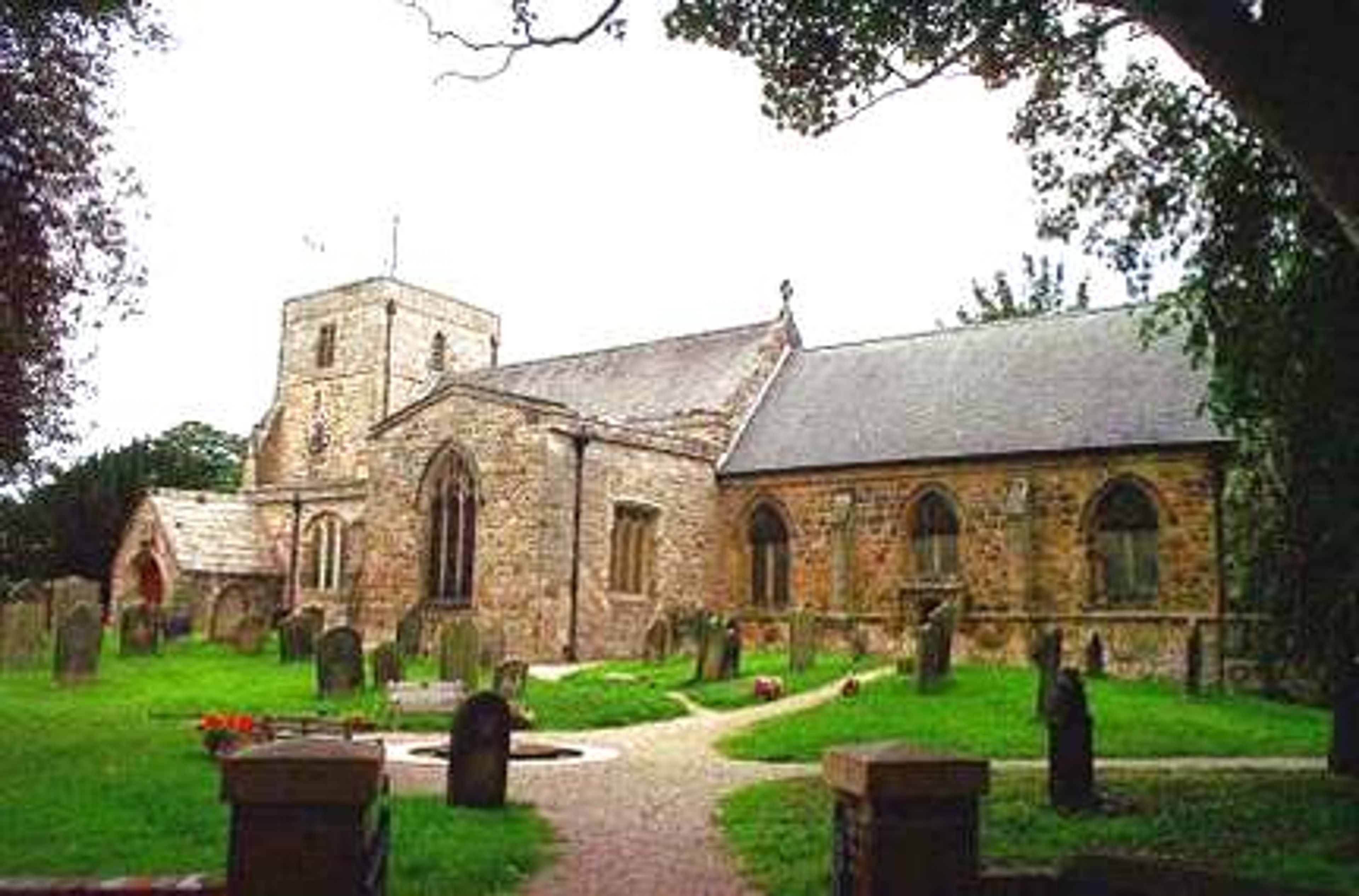 A stone parish church with a battlemented tower and arched windows, seen across a graveyard with headstones and a path