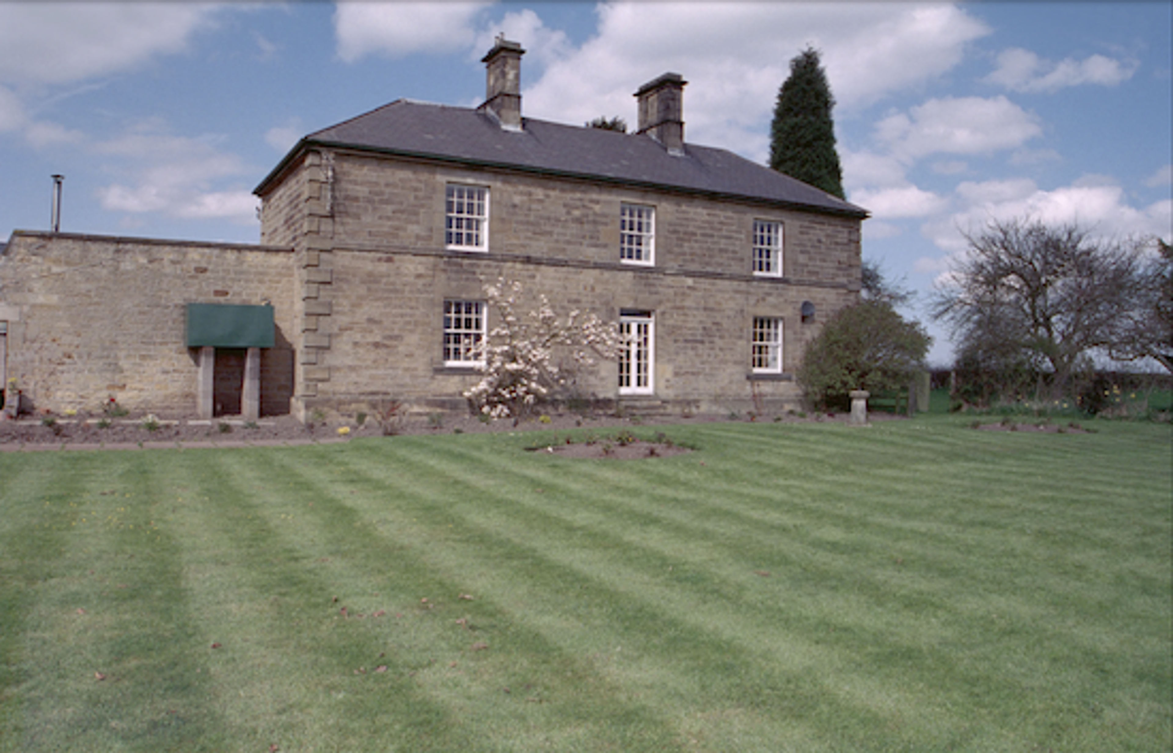 A stone farmhouse with white sash windows and two chimneys, standing behind a wide striped lawn