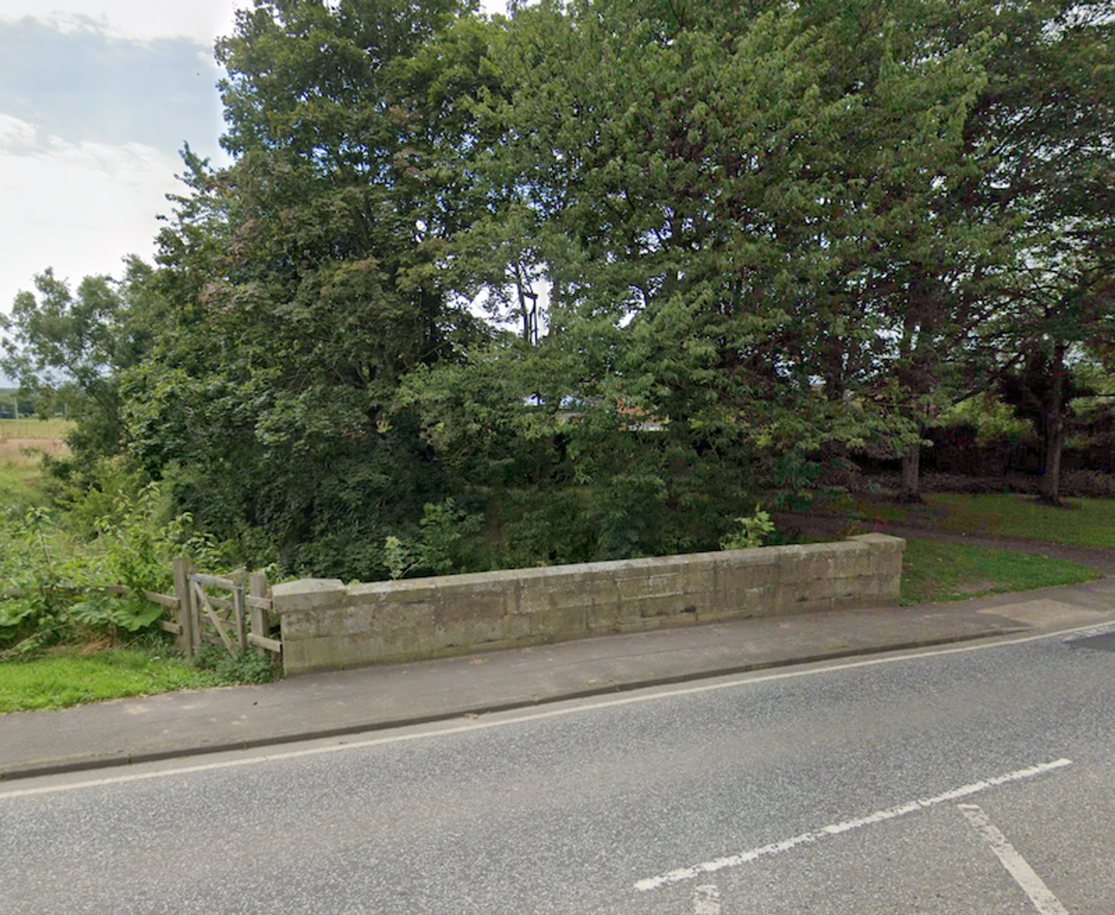 A low stone parapet bridge carrying a road, half hidden by heavy tree cover, with a field gate to one side