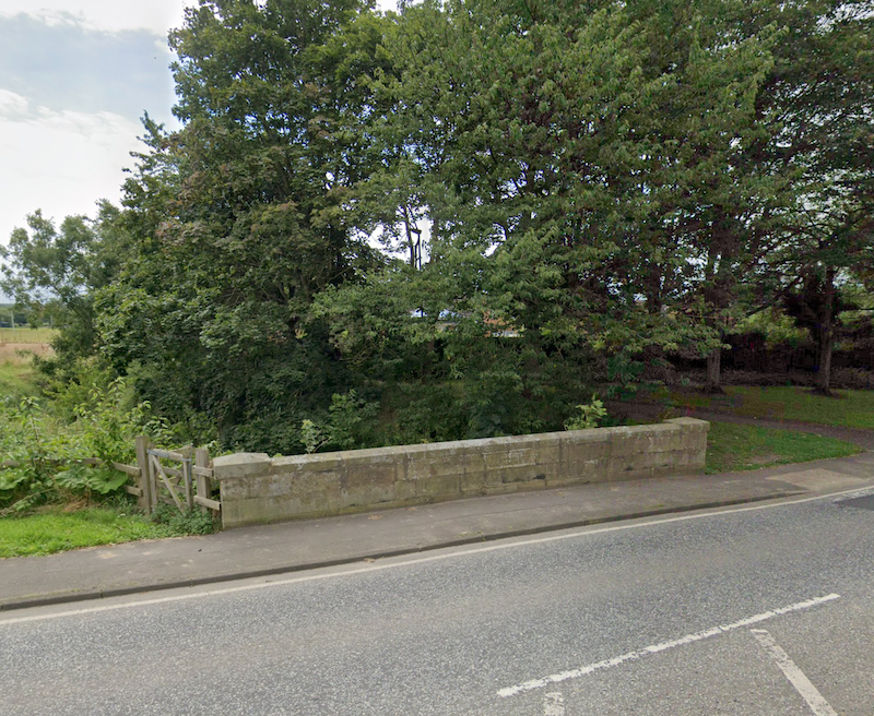 A low stone parapet bridge carrying a road, half hidden by heavy tree cover, with a field gate to one side