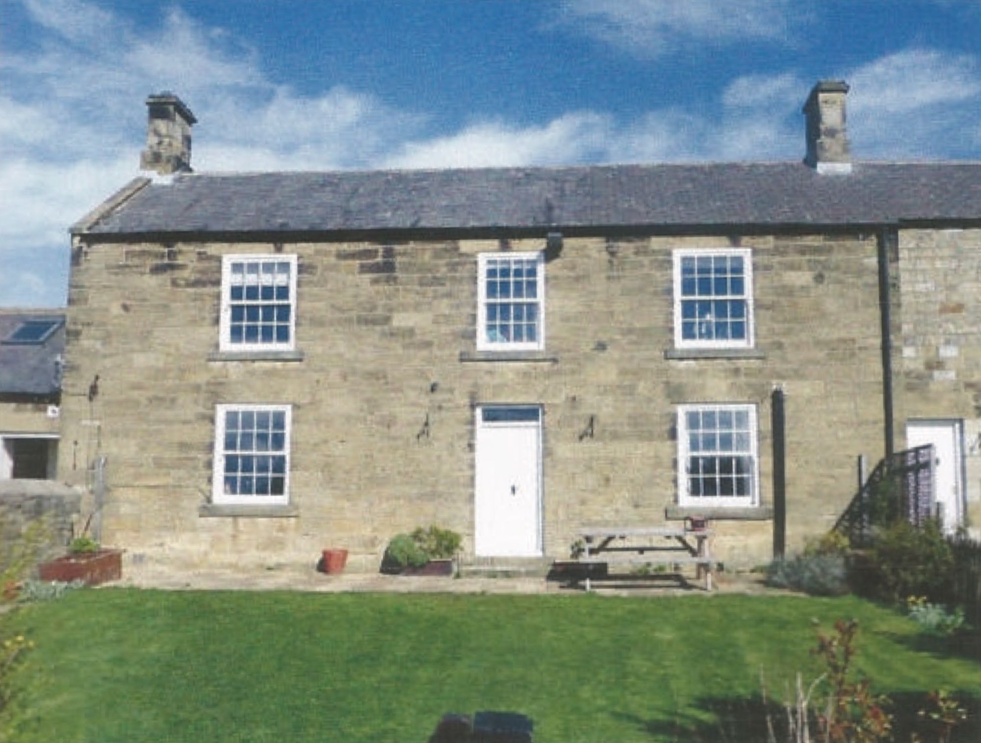 A stone farmhouse with sash windows and two chimneys, a picnic bench on the lawn in front