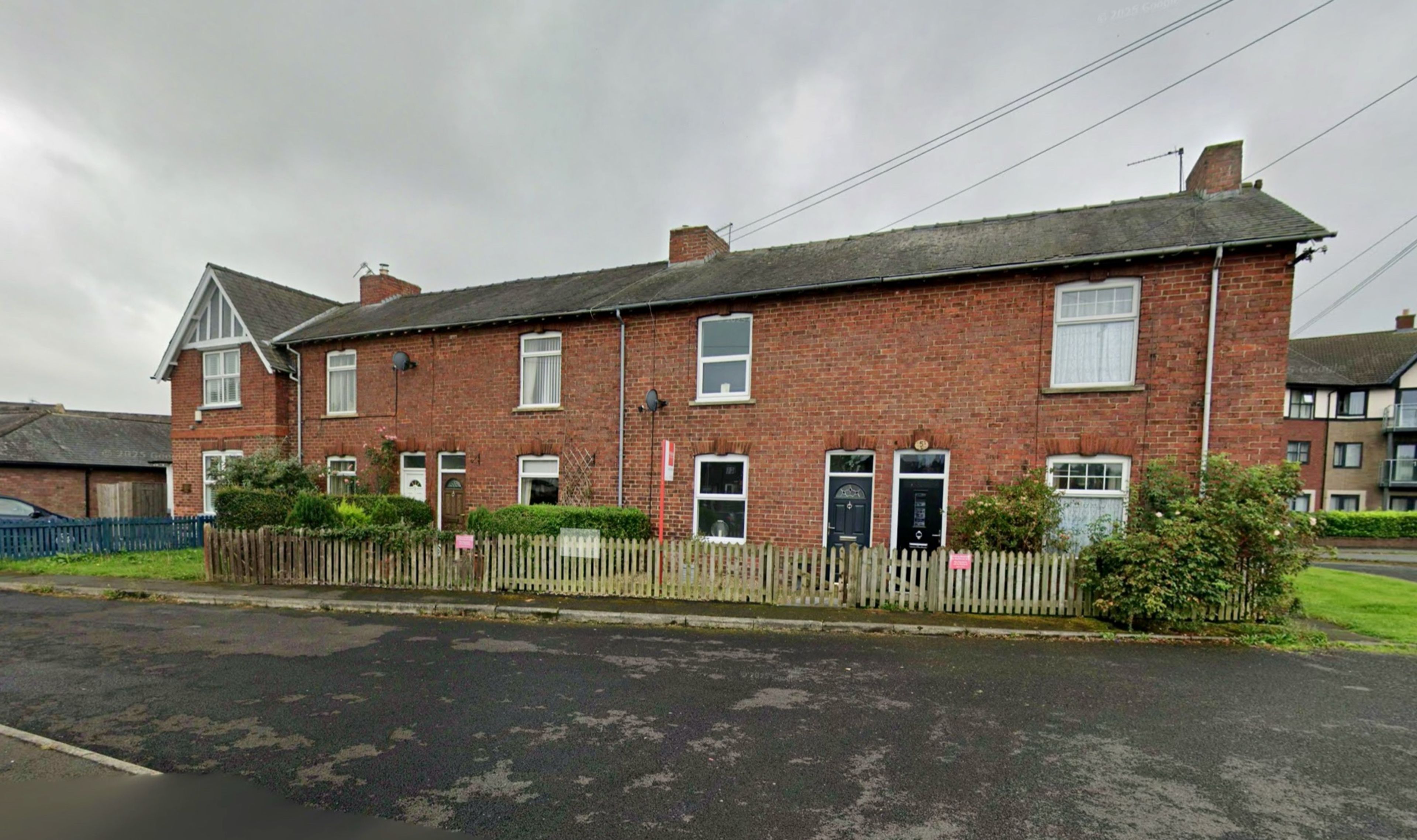 A terrace of red brick cottages with chimney stacks and small front gardens behind a wooden fence, seen from the road