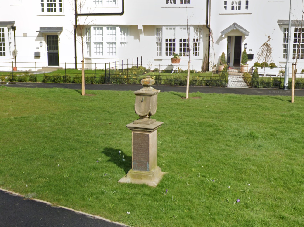 A stone pedestal sundial with a carved urn finial standing on a lawn in front of white rendered houses
