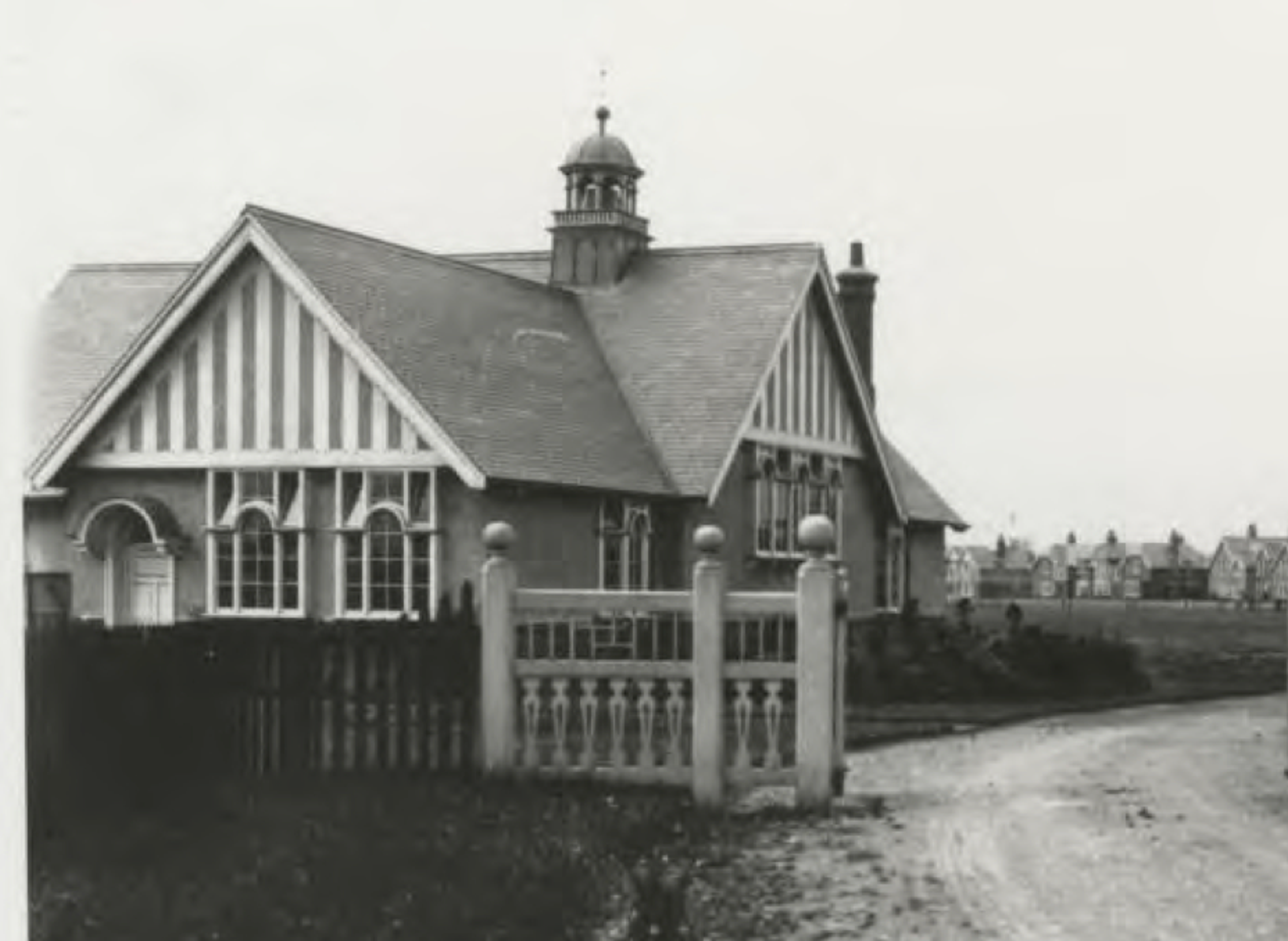Black and white photograph of a decorative gabled school house with a bell turret, behind an ornamental balustrade