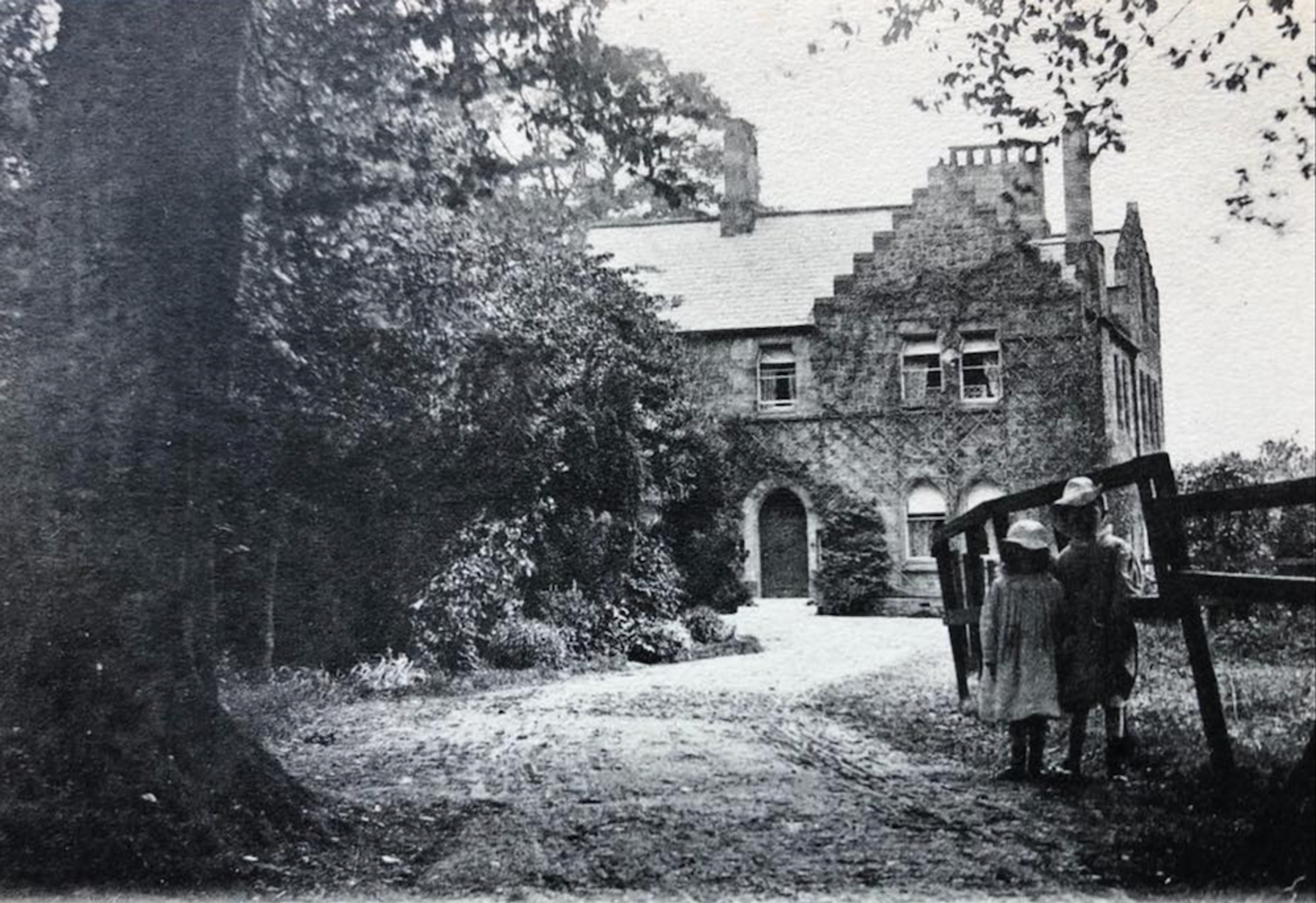 Black and white photograph of a large gabled house half hidden by trees, with two children in hats standing by a fence in the foreground