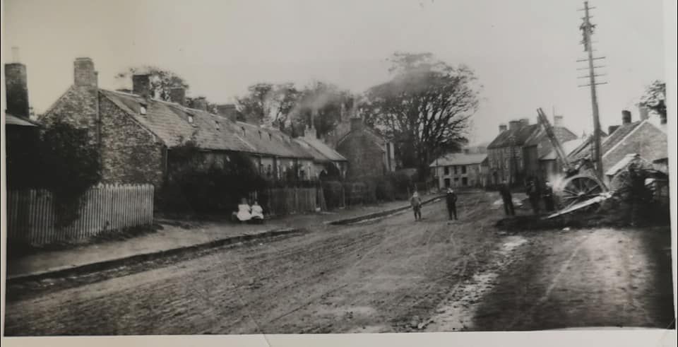 Black and white photograph of an unmade village street with stone cottages on the left, figures standing in the road and a telegraph pole