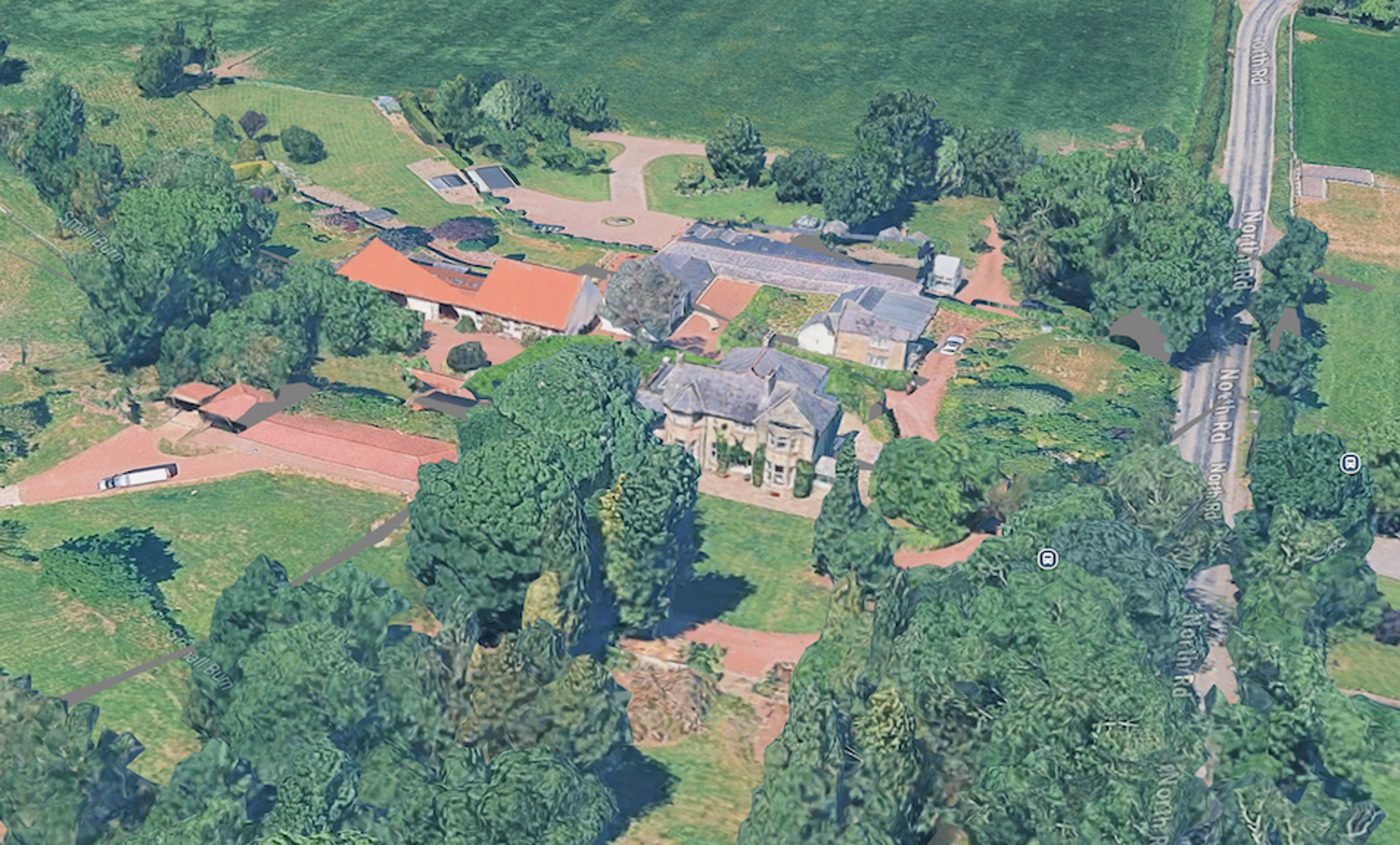 Aerial view of a house with a slate roof among mature trees, with long red-roofed farm buildings alongside