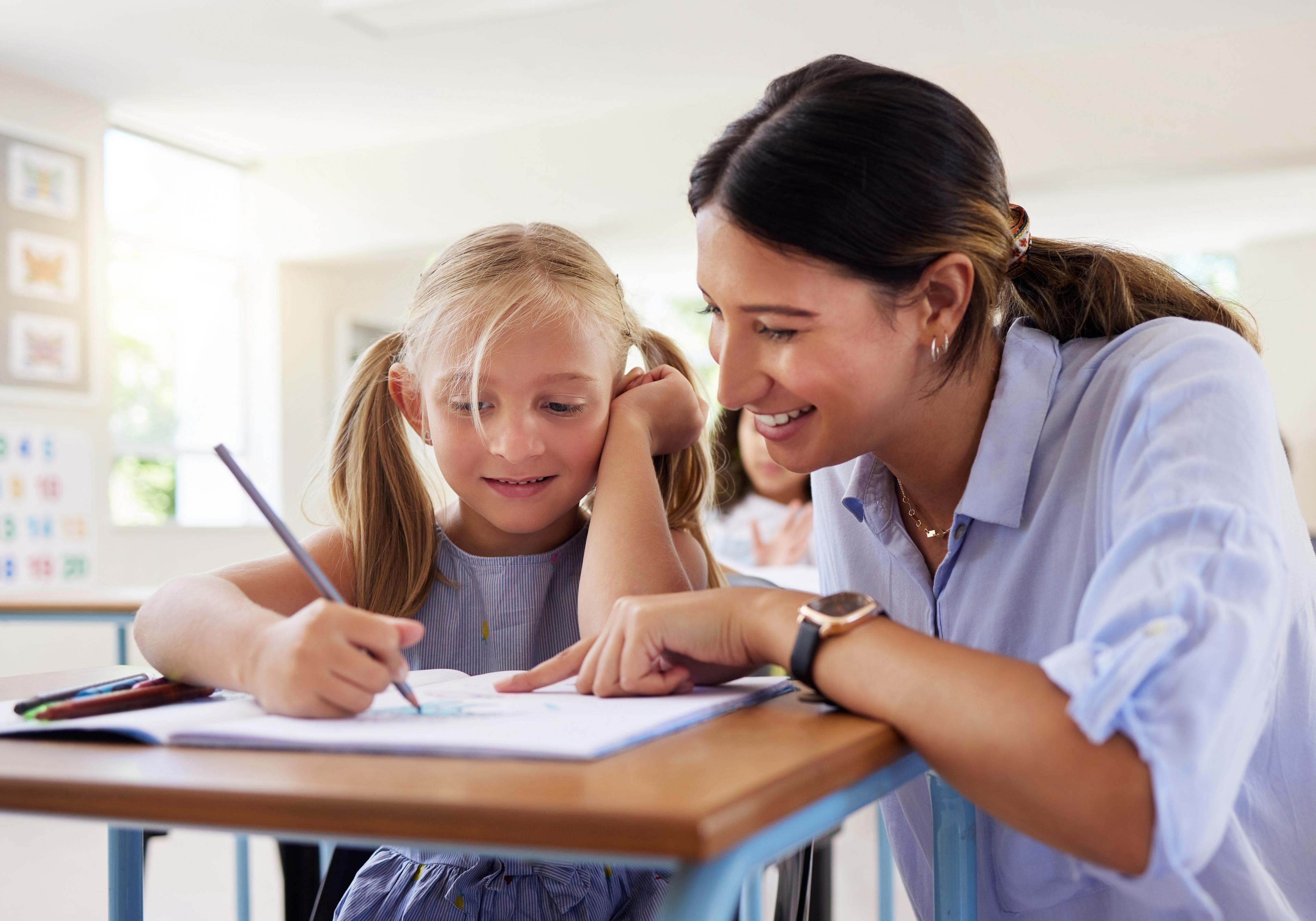 A teacher smiling as she helps a young student with her drawing at a classroom desk