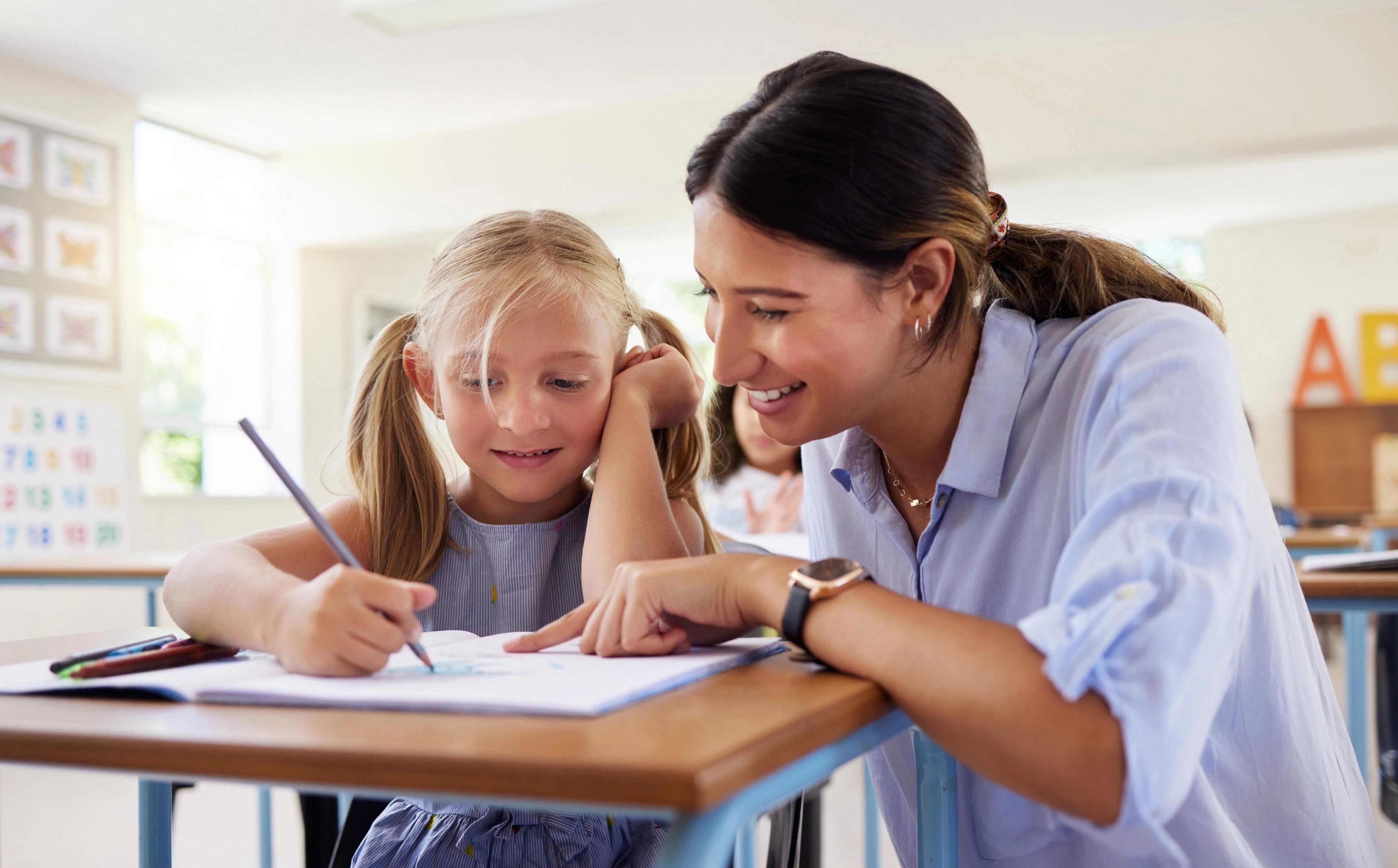 A teacher smiling as she helps a young student with her drawing at a classroom desk