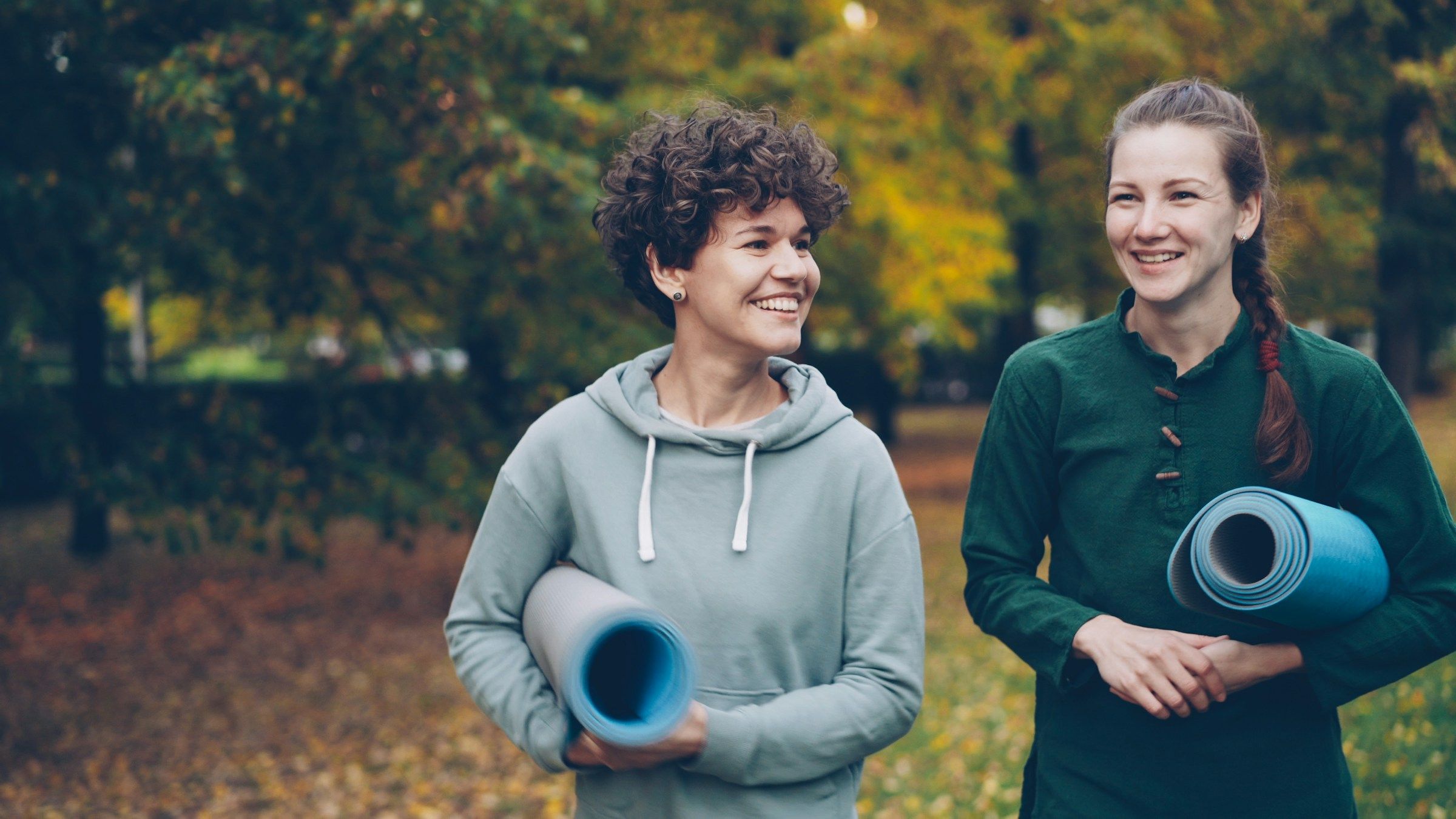 Two smiling women walking in a park carrying yoga mats