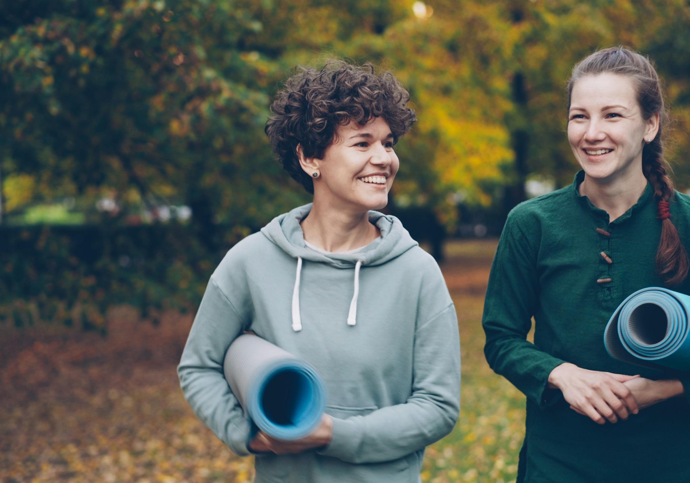 Two smiling women walking in a park carrying yoga mats