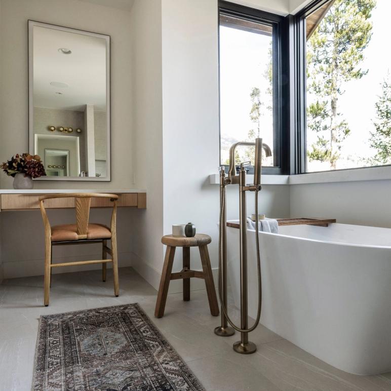 Bathroom with a freestanding white tub under a corner window onto pine trees, a brass floor-standing filler, and a built-in oak vanity