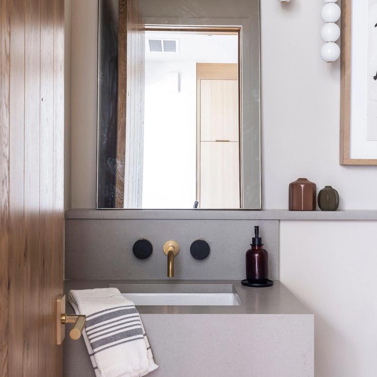 Powder room with a floating grey stone basin, wall-mounted brass tap and an arched mirror, beside a white-oak door