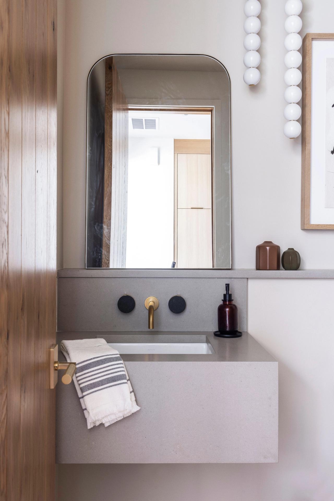 Powder room with a floating grey stone basin, wall-mounted brass tap and an arched mirror, beside a white-oak door