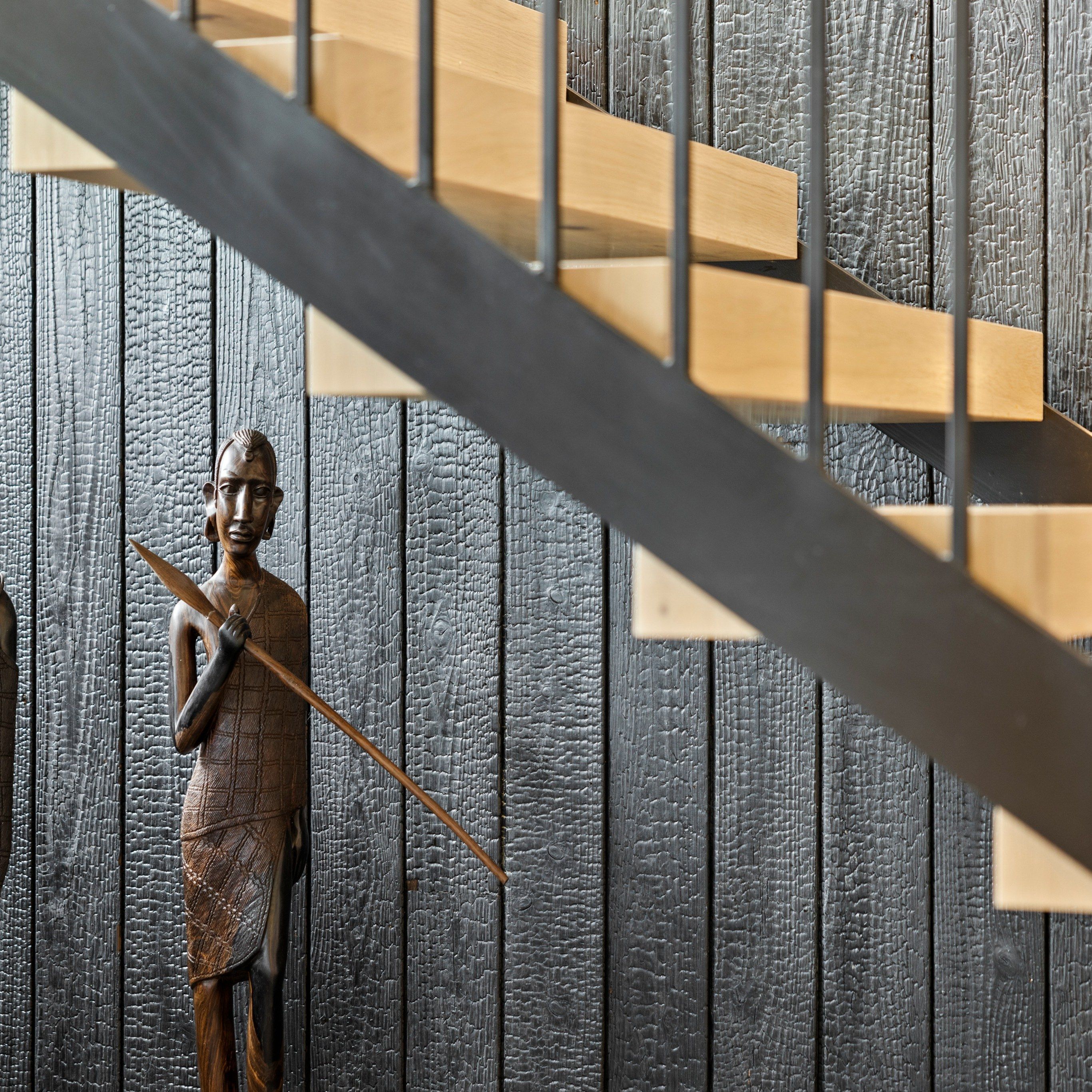 Carved wooden figure standing against charred-timber cladding, beneath the floating oak treads and black steel of a staircase