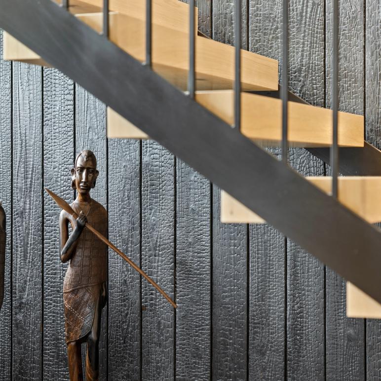Carved wooden figure standing against charred-timber cladding, beneath the floating oak treads and black steel of a staircase