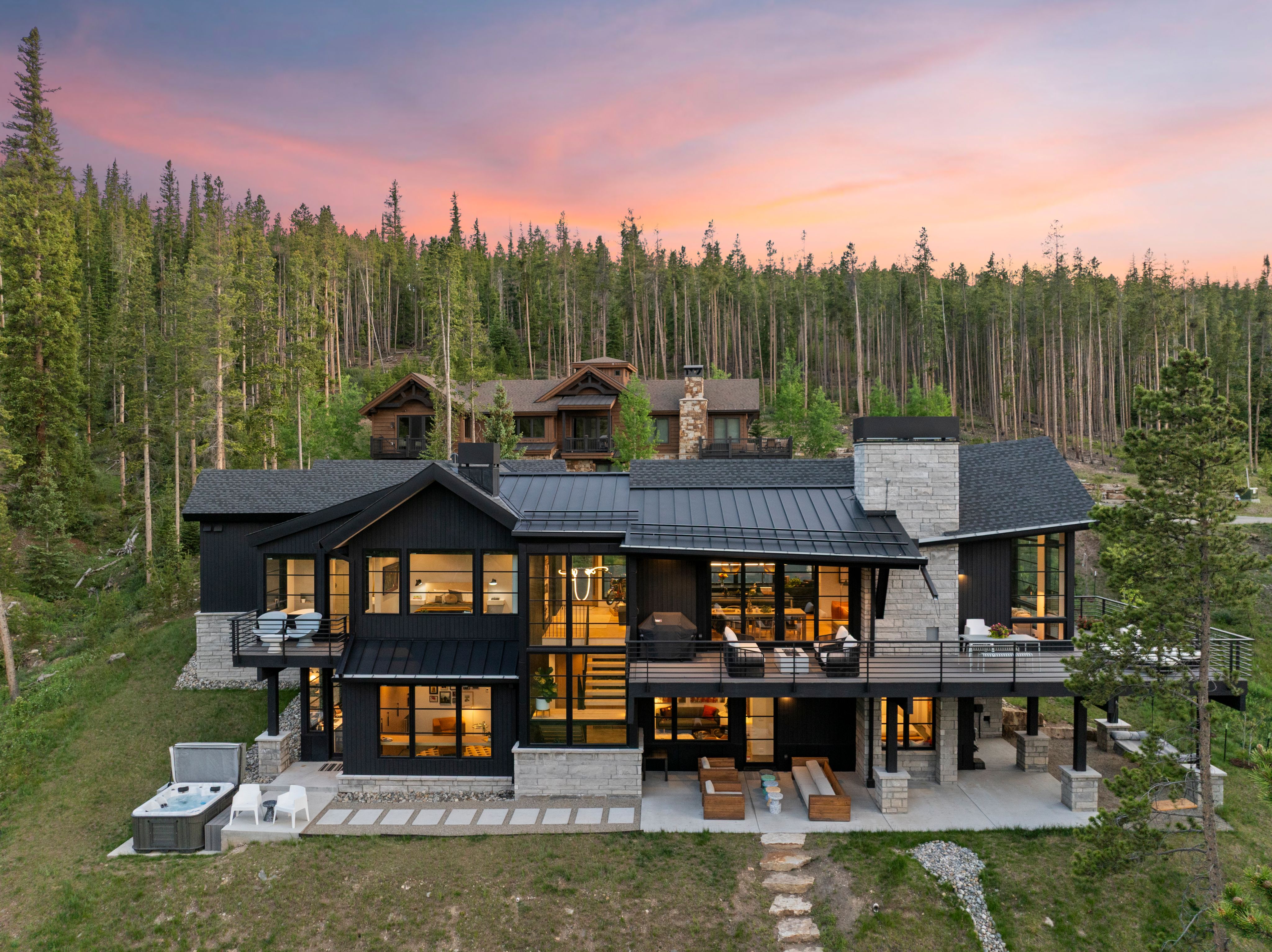 Modern Iron Forest mountain home at dusk, dark siding and stone chimney against a Breckenridge forest hillside