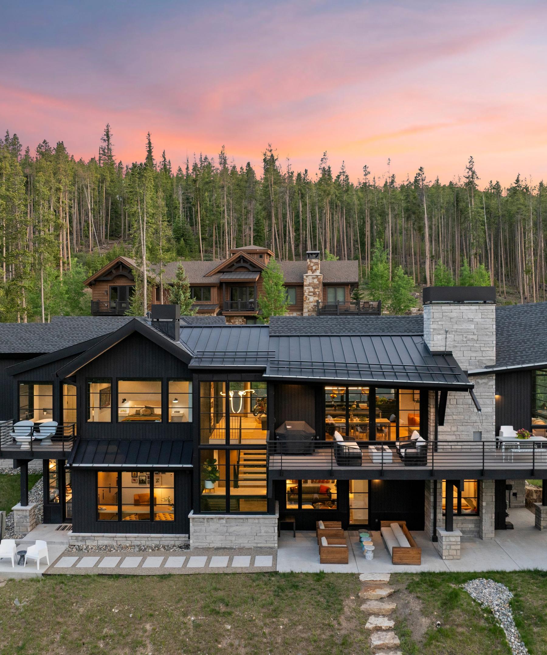 Modern Iron Forest mountain home at dusk, dark siding and stone chimney against a Breckenridge forest hillside