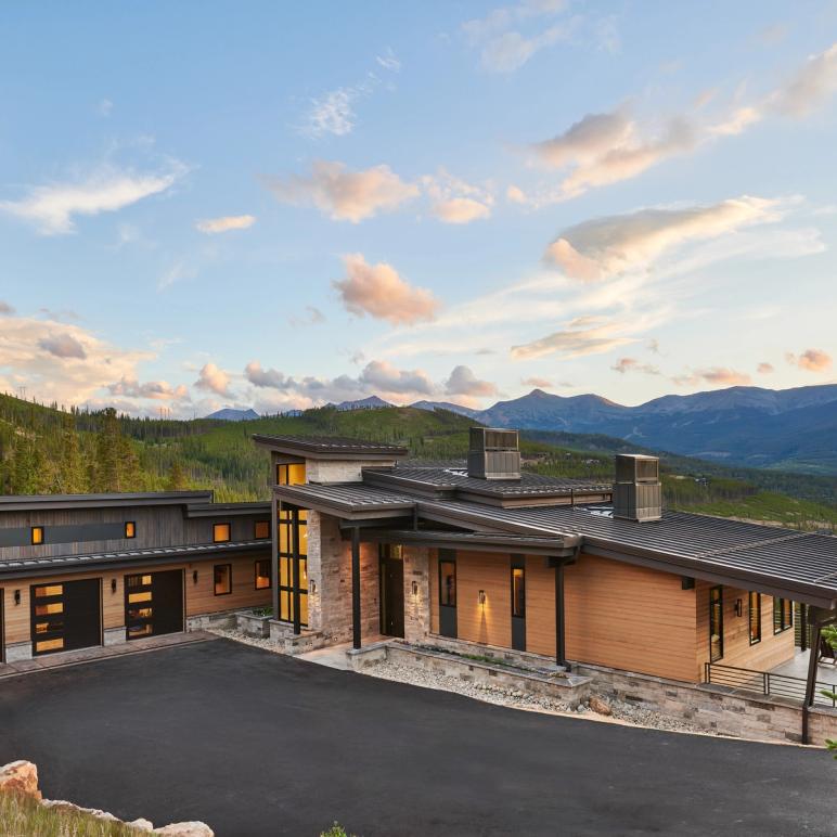 Mountain home at dusk in dark cedar, stone and standing-seam steel, windows lit, with the Tenmile Range behind