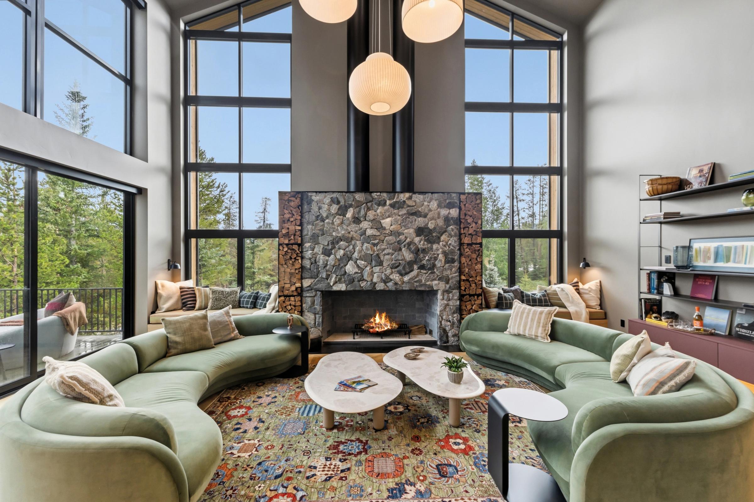 Great room with curved green sofas around a river-stone fireplace, walls of windows on both sides