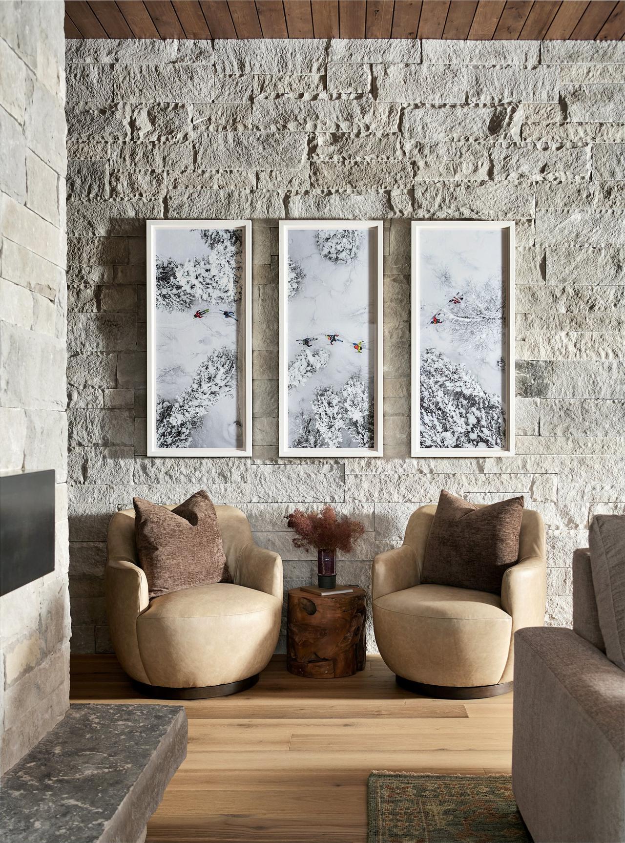 Sitting area with two leather swivel chairs and a live-edge stump table before a stone wall hung with three aerial photographs of ski tracks