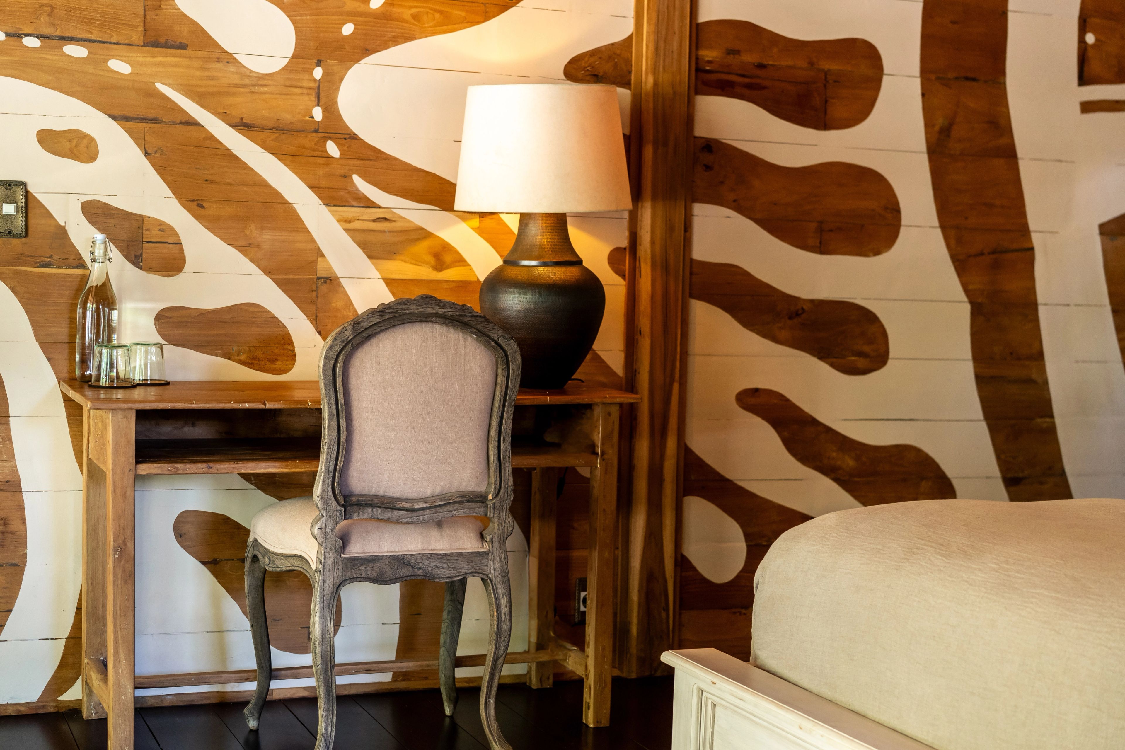 A carved chair at a dressing table with a lamp, against a wall painted in bold pale shapes.
