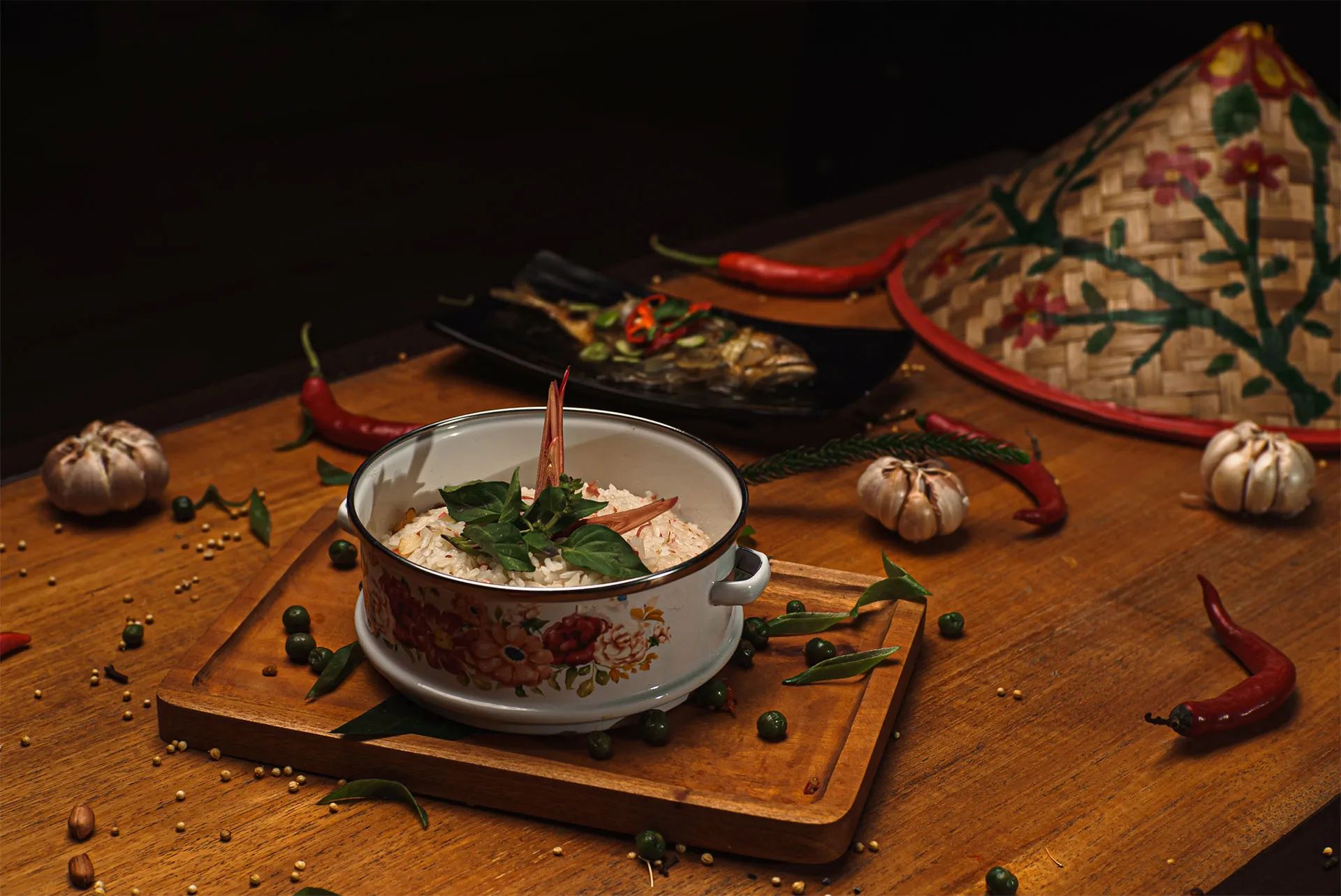 A pot of soup with chillies, garlic and herbs laid out around it on a timber board, a woven hat behind.