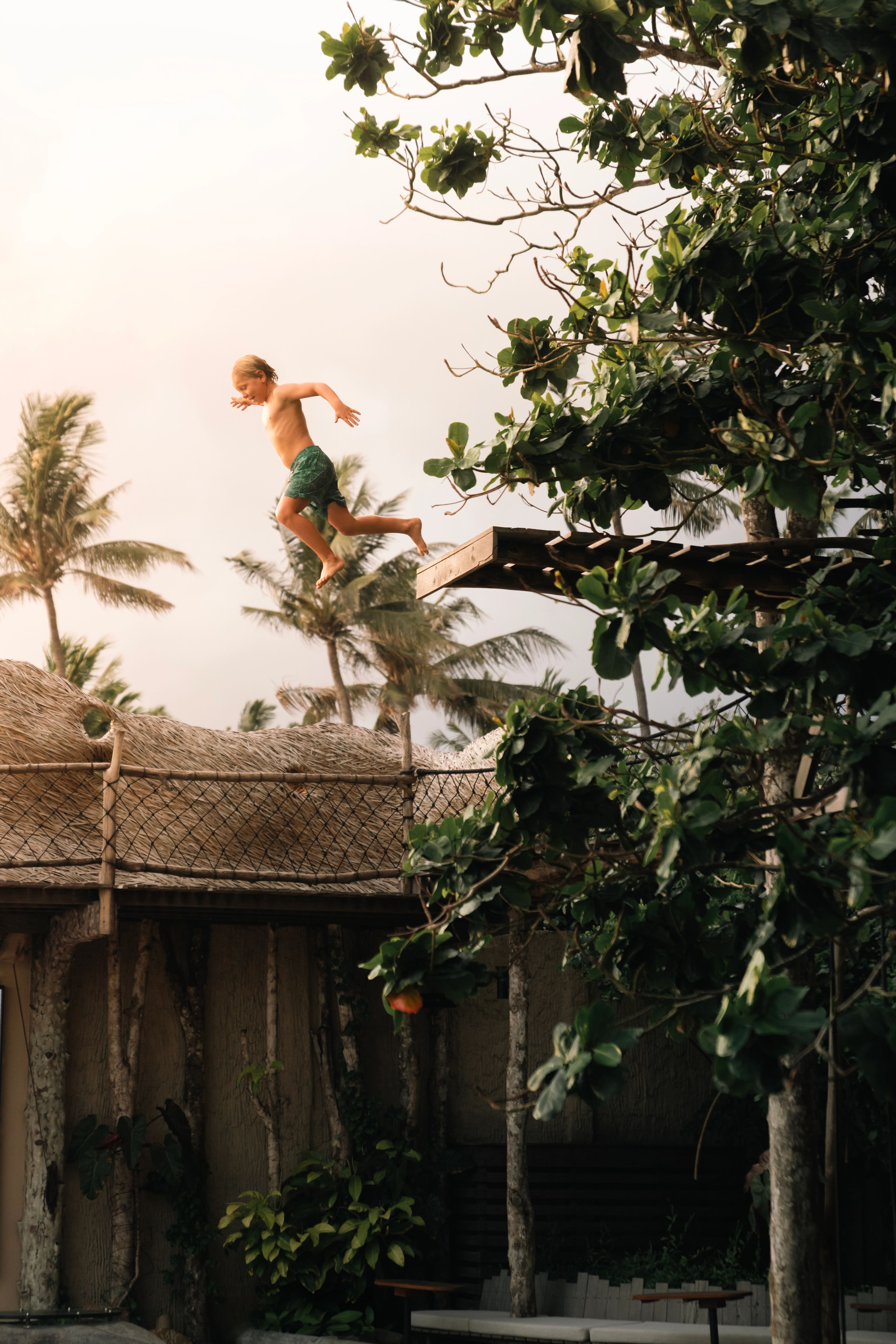 A child leaping off the timber platform above the treehouse roof, palms behind.