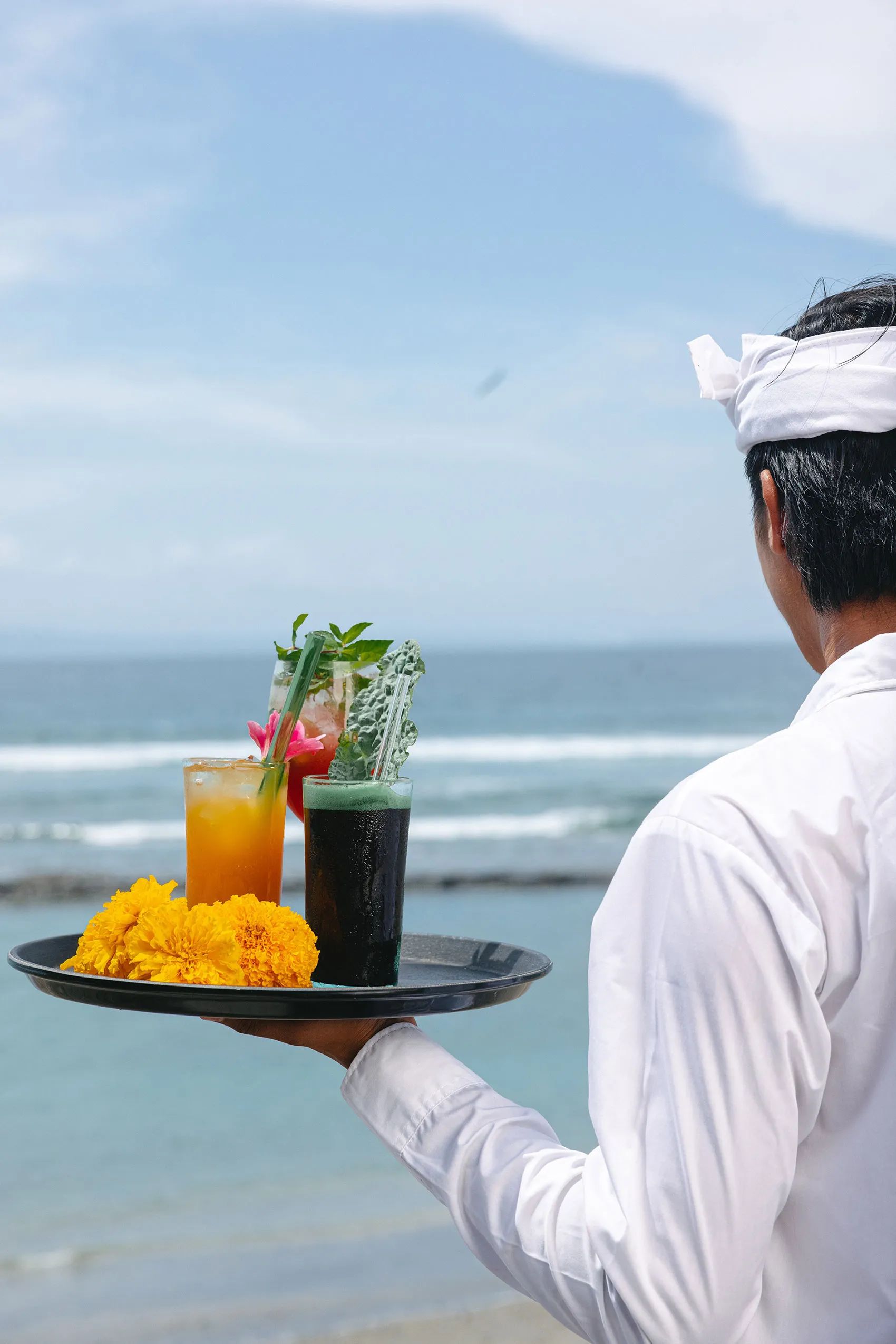 Two cocktails garnished with flowers and herbs on a tray beside marigolds, a member of staff carrying them down to the sea.